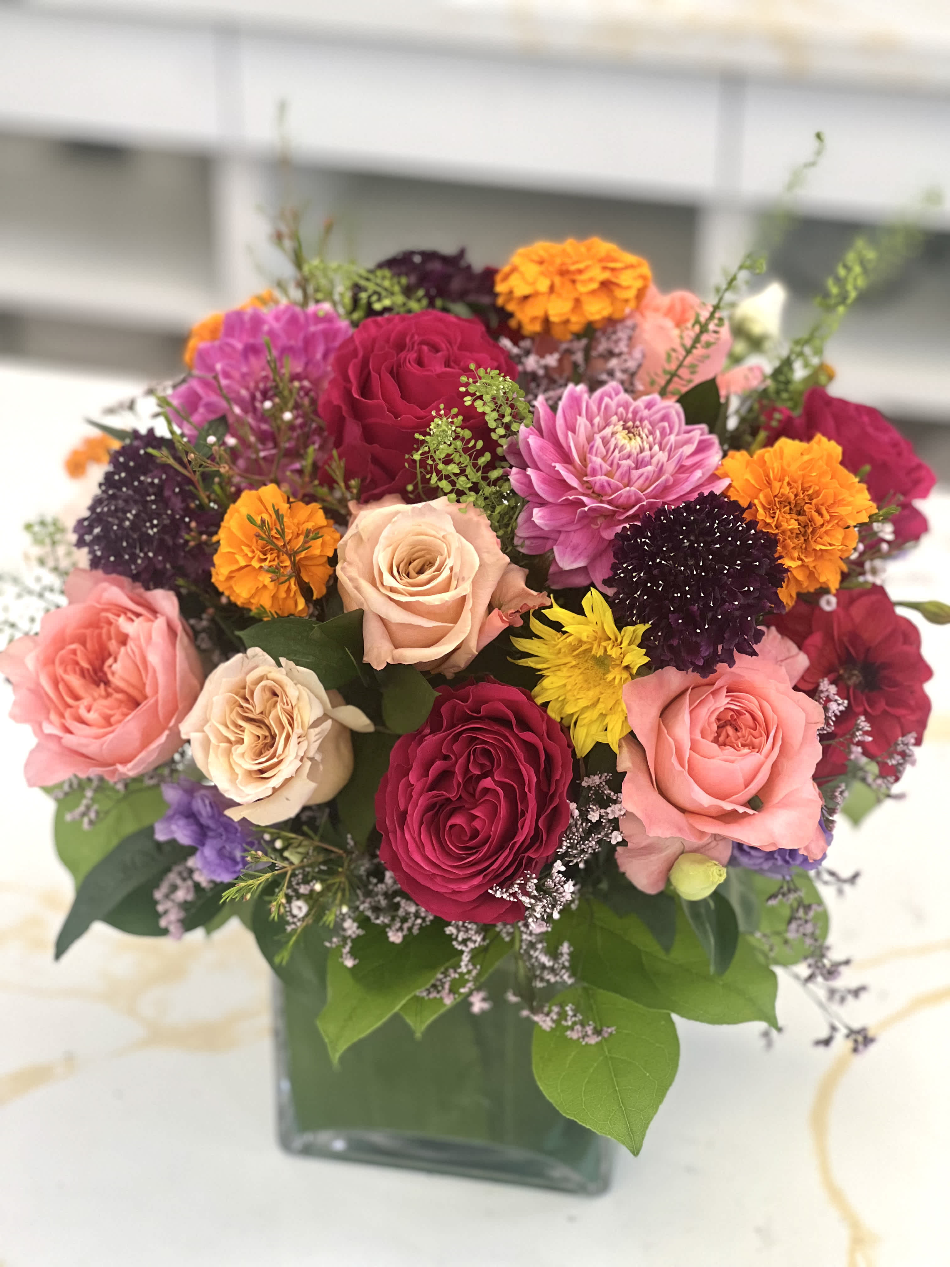Mixed bouquet of roses, dahlias, and marigolds in a square glass vase