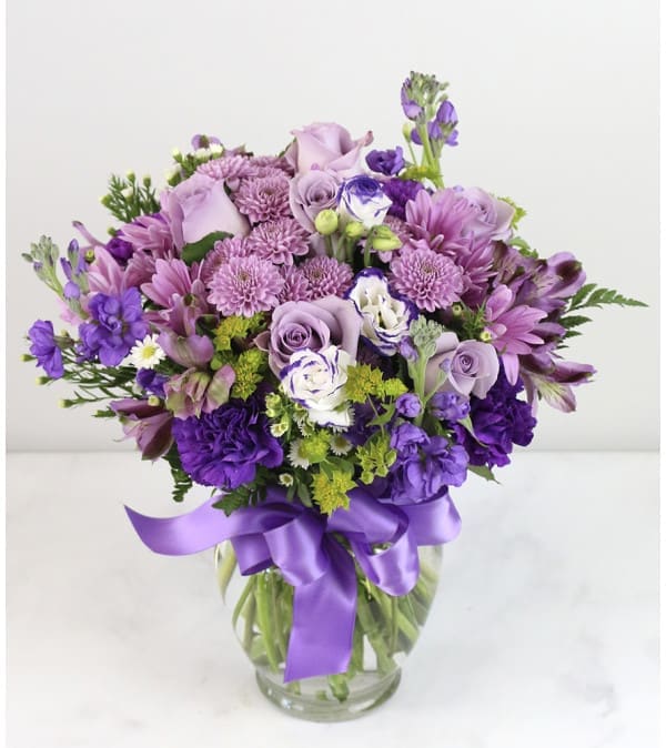 Lavender and purple mixed flower arrangement with roses and mums in a clear vase with a satin ribbon bow