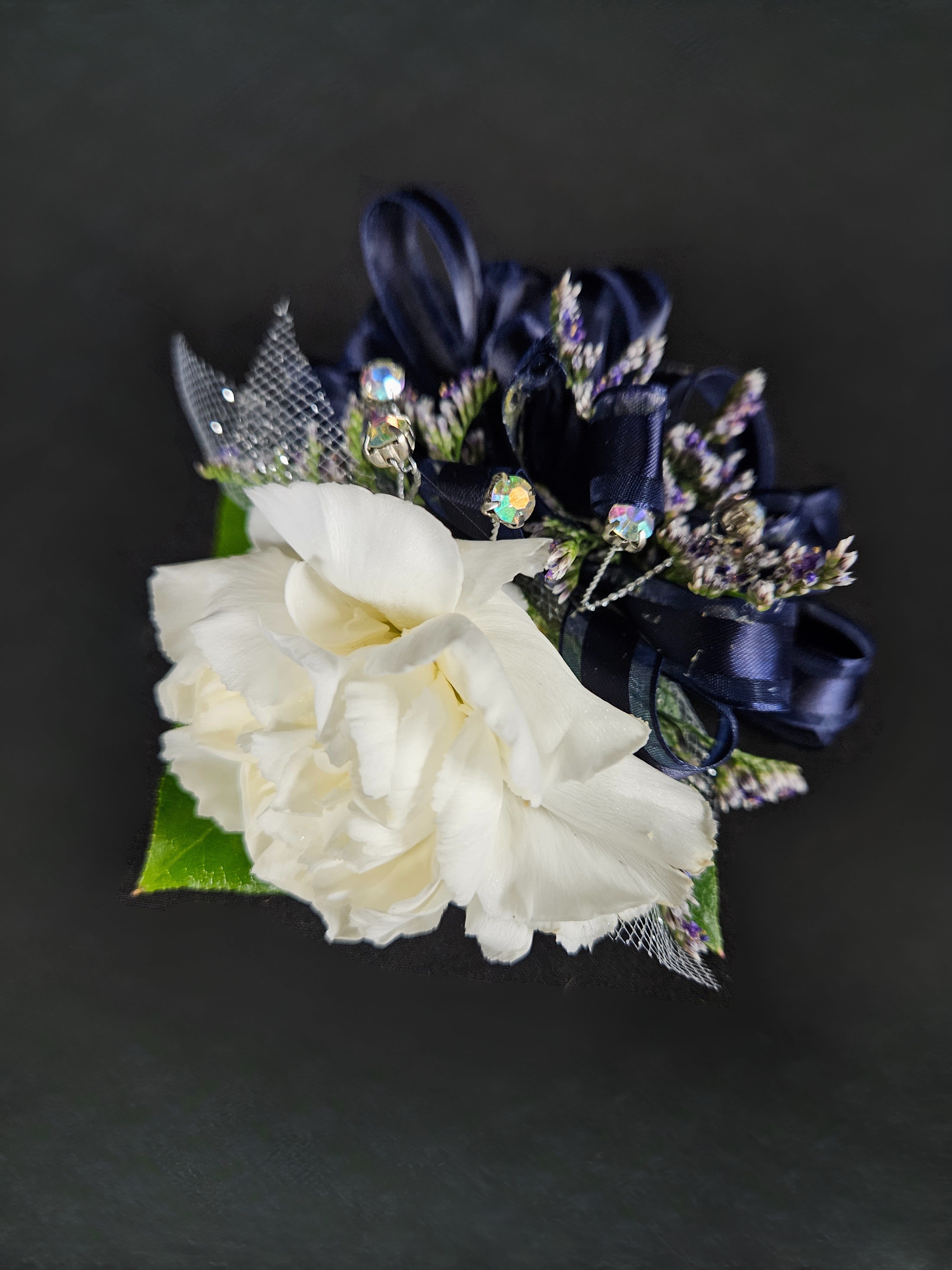 White carnation corsage with navy ribbon loops, tiny purple accents, and jeweled picks on a dark background.