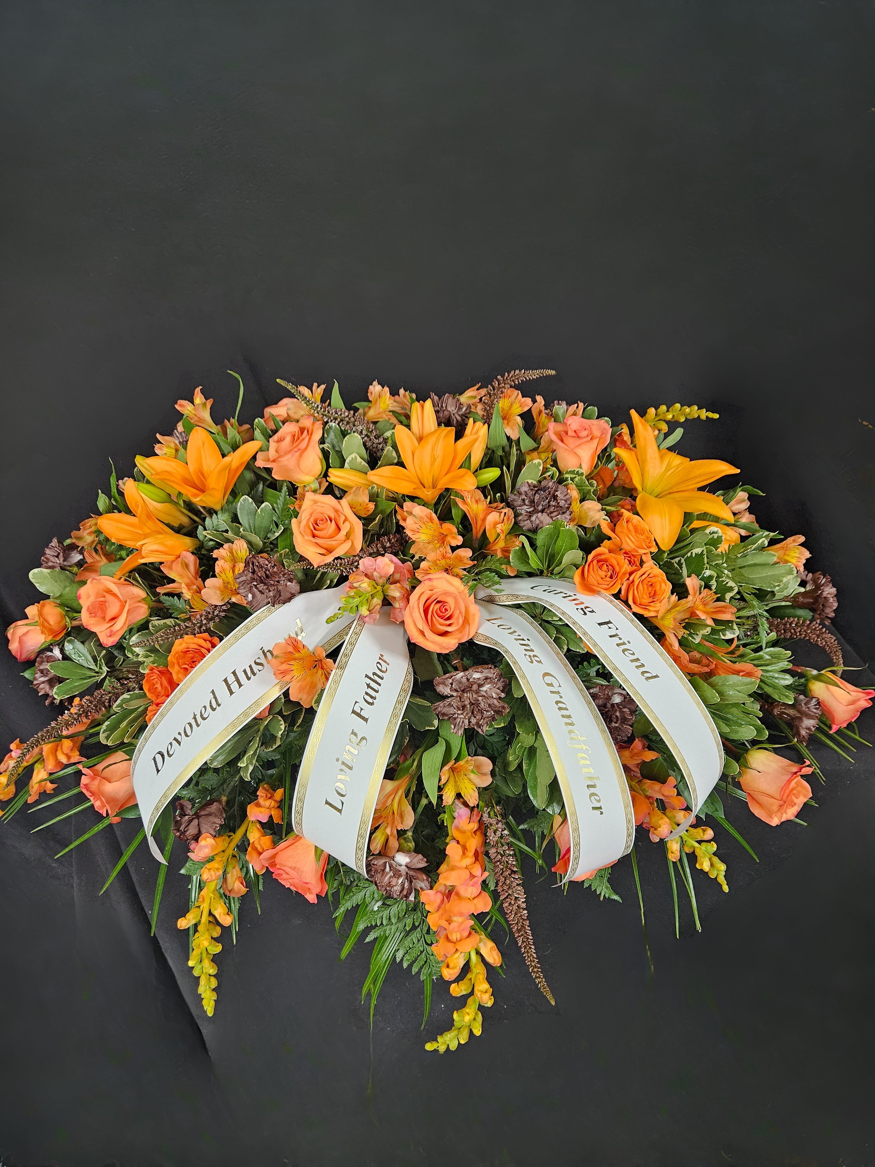 Orange lilies and roses casket spray with white sympathy ribbons on a dark background