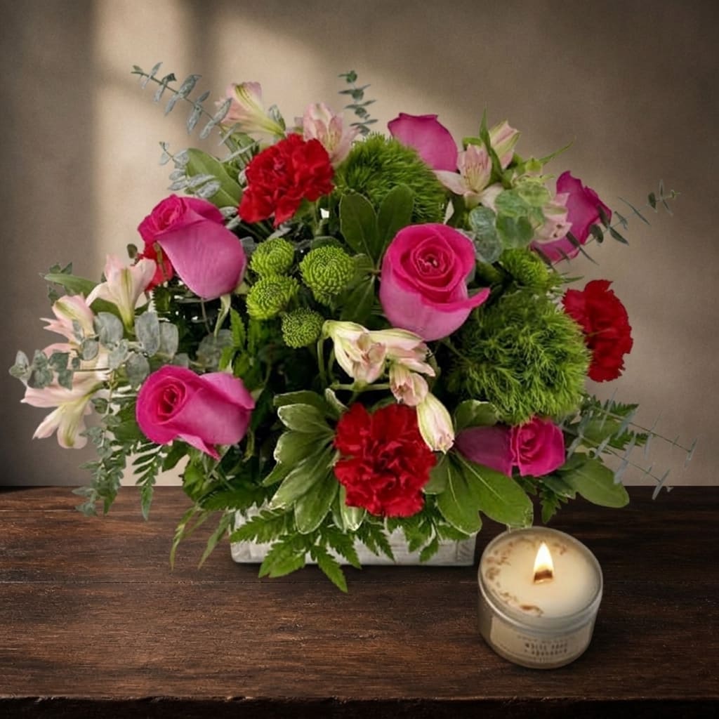 Pink roses and red carnations arranged in a low square vase with a candle nearby.