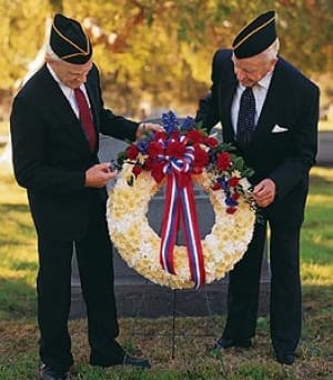 Patriotic standing wreath of white flowers with red, white, and blue accents and ribbon on an easel outdoors.