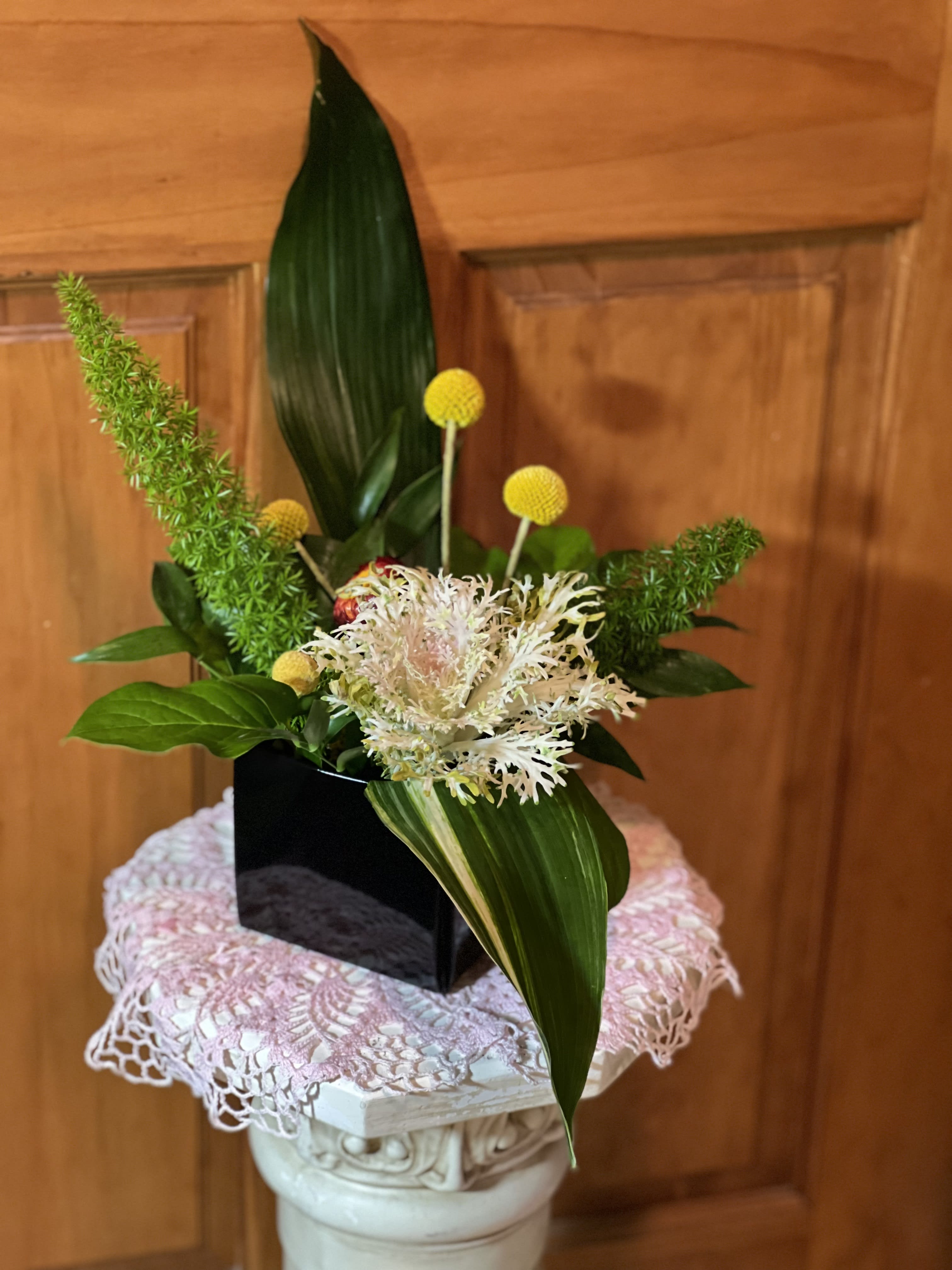 Compact floral arrangement with white blooms, yellow accents, and broad green leaves in a black vase