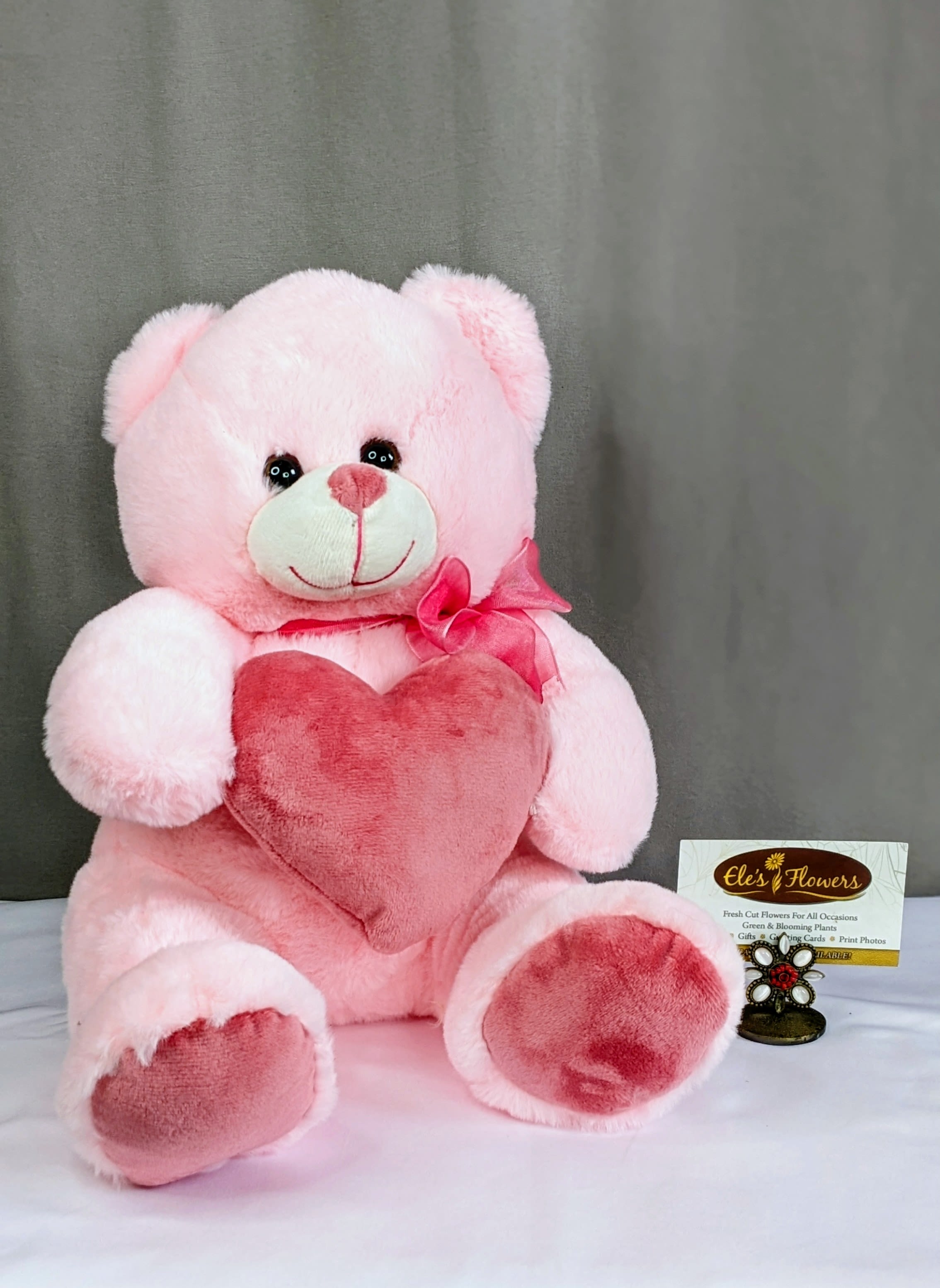 Pink teddy bear plush holding a darker pink heart pillow with a bow at its neck on a neutral backdrop