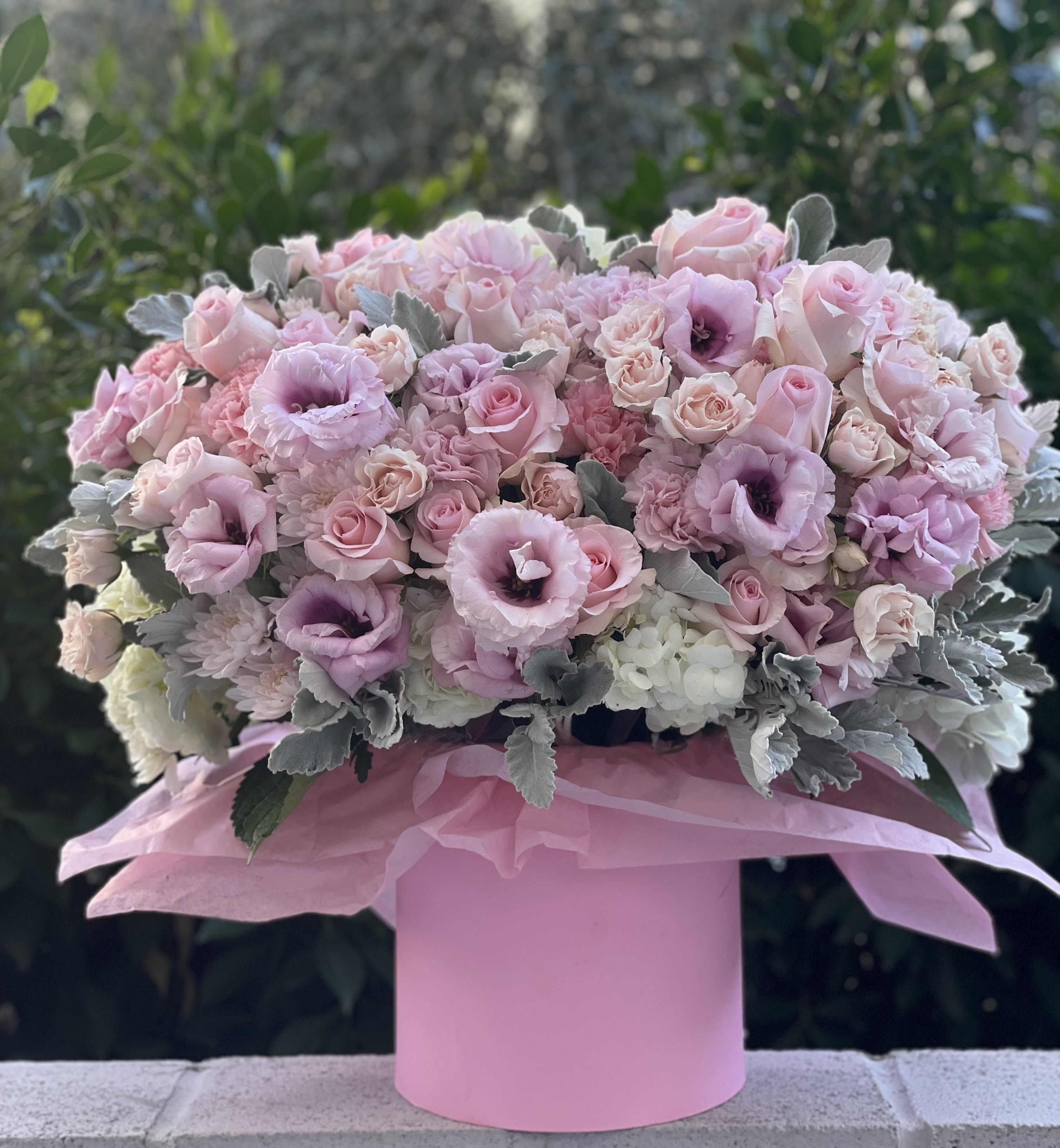 Pink dreams box - A plush dome of blush and lavender florals artfully arranged in a pink round hatbox-this flower arrangement for birthdays, anniversaries, Mother's Day, new babies, or a gentle "thinking of you" moment reads as tender and celebratory the instant she sees it. The Pink dreams box is an ideal choice for romantic gestures, get-well wishes, graduations, or as an elegant sympathy or thank-you gift that feels personal and thoughtful.  Tightly massed roses, spray roses, lisianthus, ruffled carnations, ranunculus, chrysanthemums, and creamy white hydrangeas form a cloud-like, feminine bouquet with soft silvery-green foliage peeking through for contrast. The blooms are finished with layered pink tissue paper that cushions the stems and spills slightly over the matte pink hatbox, creating a polished presentation that sits beautifully on a bedside table, mantle, or party gift table. Size and care: delivered in a water source within the hatbox; refresh water source as directed and keep in a cool spot out of direct sunlight for longest life.  Perfect for seasonal gifting from spring through winter-use it for holidays like Christmas or Mother's Day, milestone moments like graduations and anniversaries, or small mercies like "I'm sorry" and "just because" surprises.