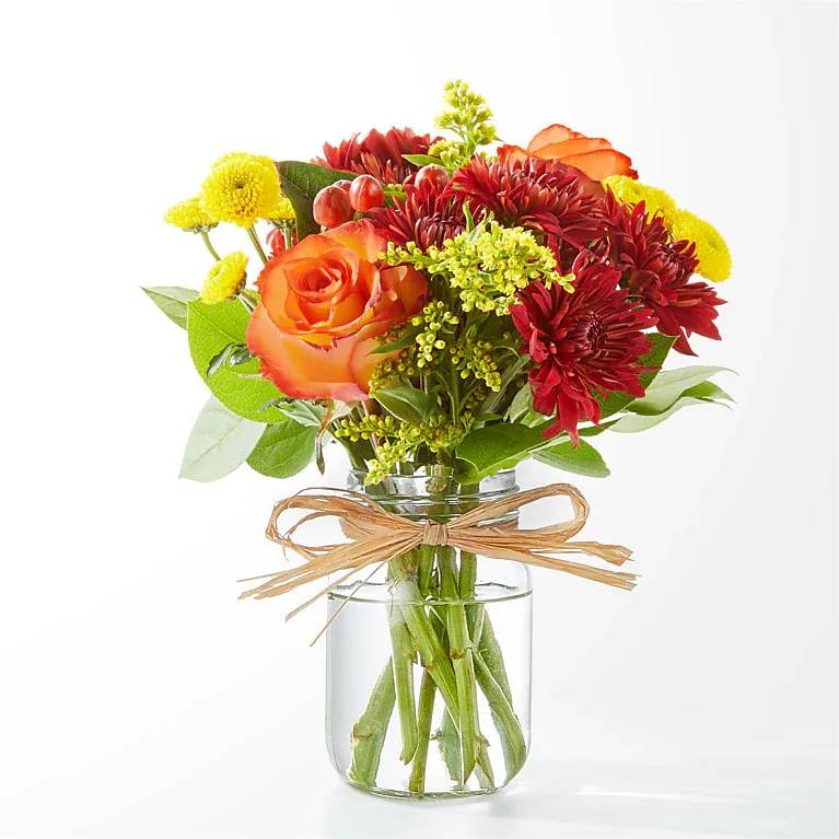 Orange and red mixed bouquet in a clear glass jar vase with a raffia bow