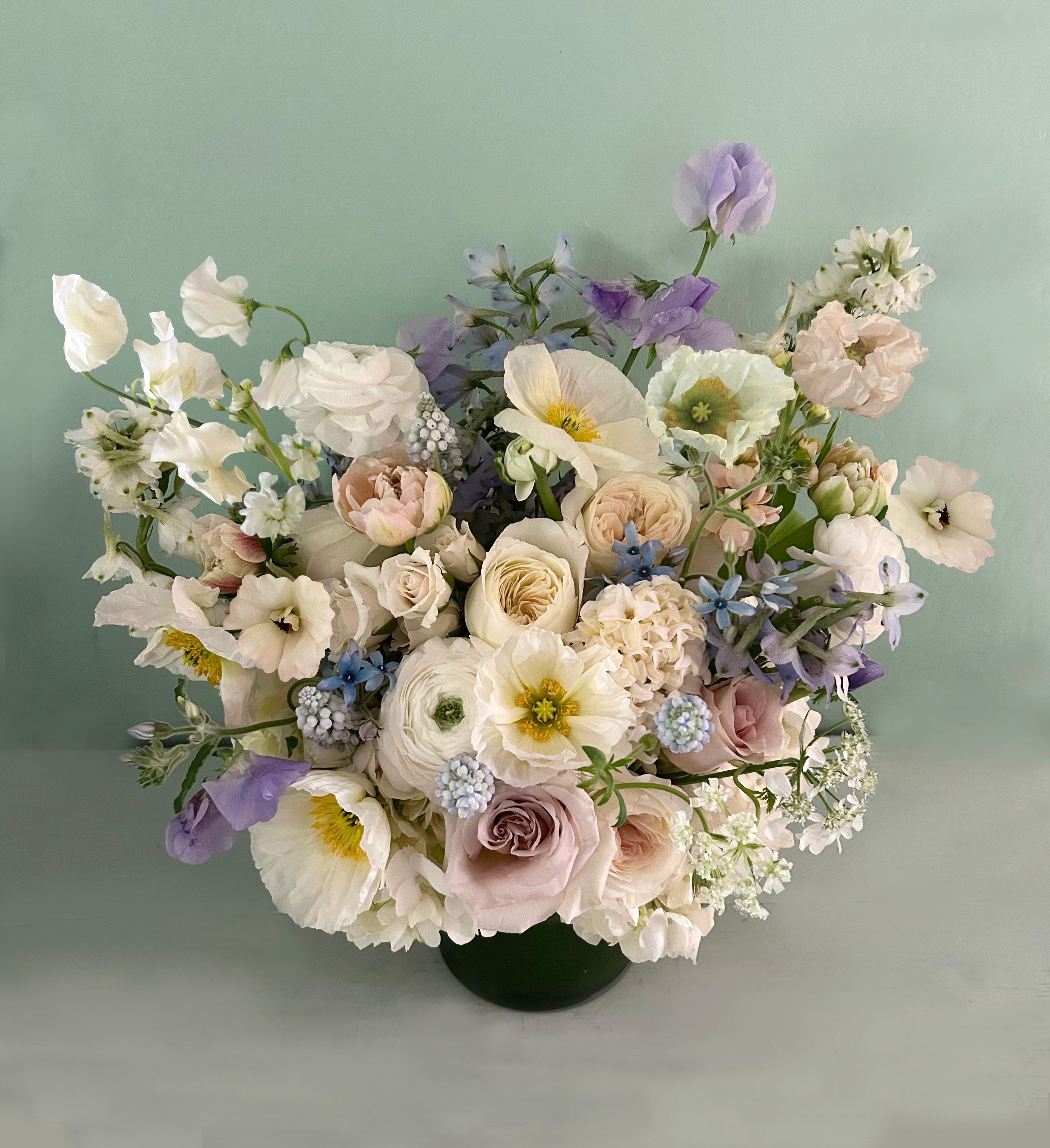 The Bette Blues - Stunning beauty in this luxe, large arrangement in muted colors of dusty blue, white and ivory colors. Named after Hollywood's Bette Davis and her striking, large blue eyes. Placed in a clear cylinder vase 6" by 6" with stems that mesmerize and capture the senses. Poppies, ranunculus, hyacinths (at winter time, cosmos at spring/summer time), garden roses with scent and other luxe seasonal stems is used to create maximum beauty and elegance. Ready for hand deliver in LA for occasions like love, sympathy, birth of a child or hannukah.