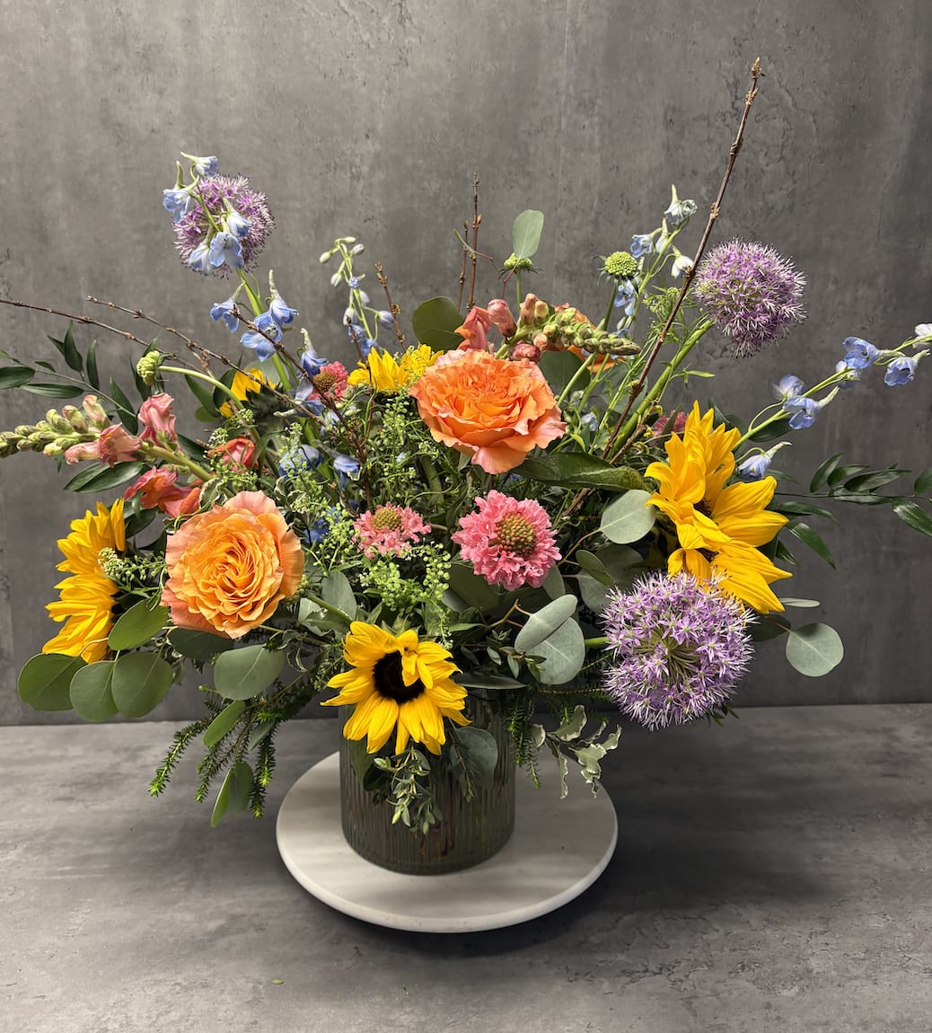 Mixed bouquet with orange roses, sunflowers, and purple allium in a glass vase