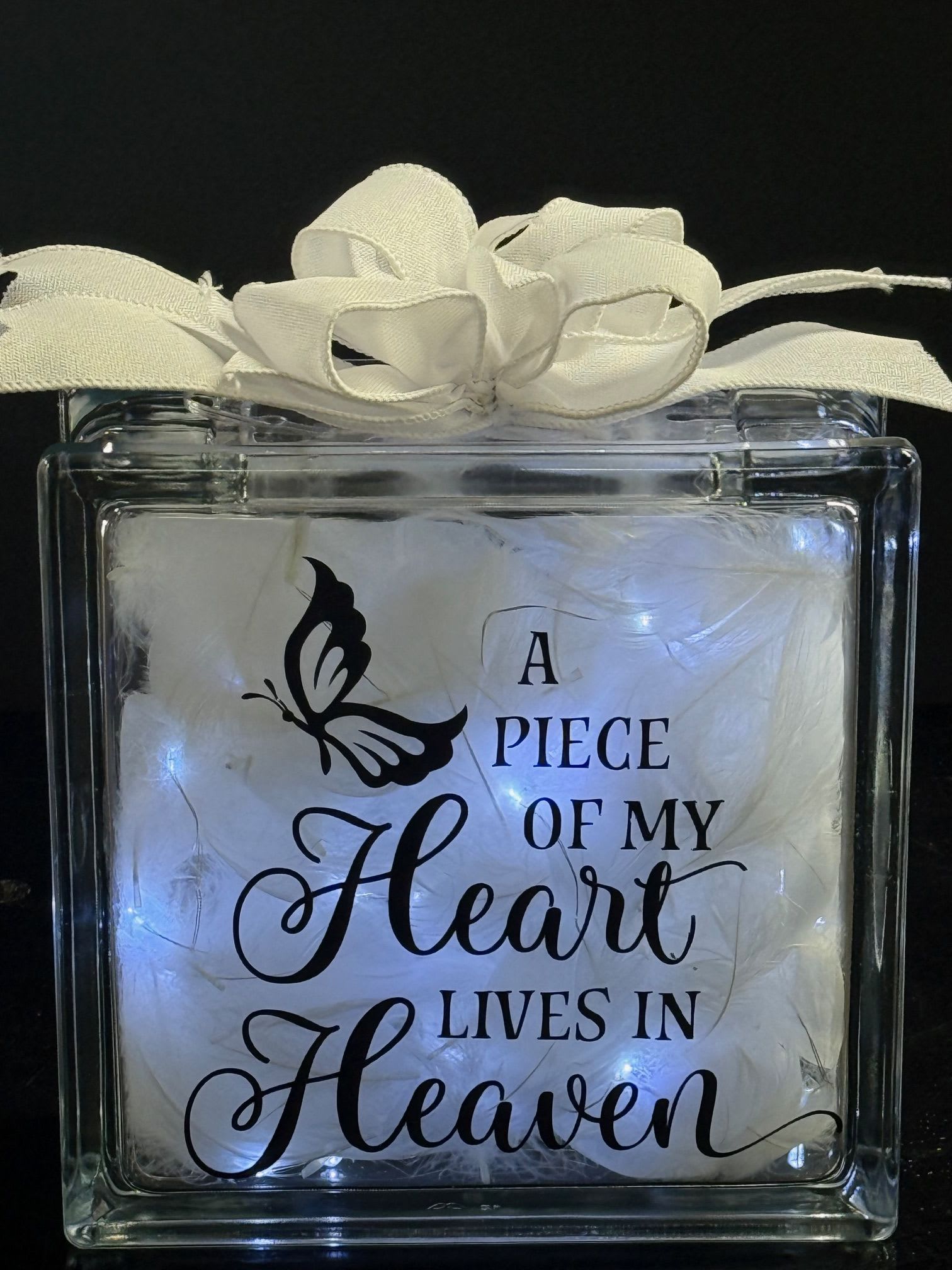 Glass block light with white bow, feathers, and memorial text 'A piece of my heart lives in Heaven'.