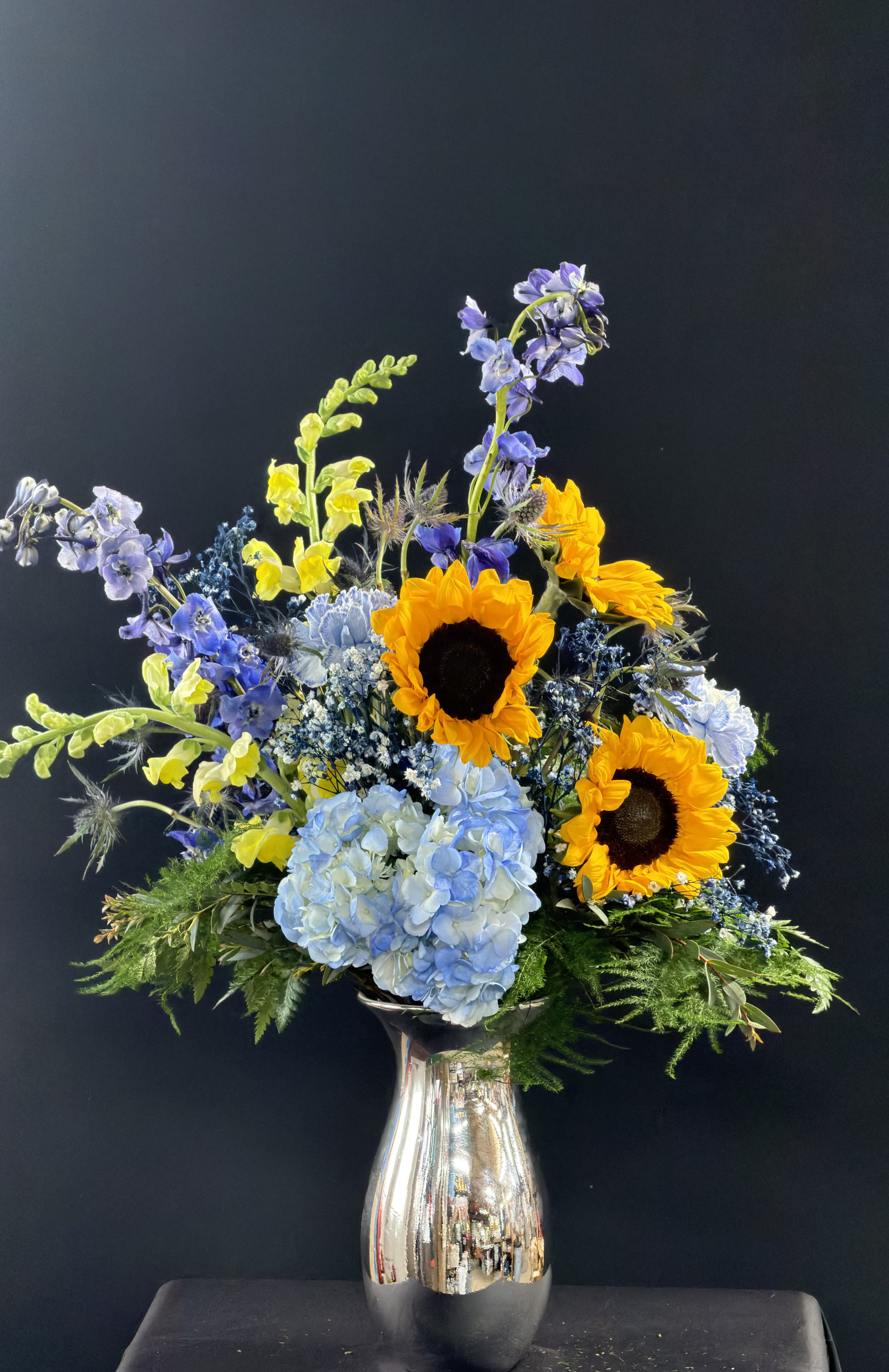 Mixed bouquet with sunflowers, blue hydrangea, and purple flowers in a silver vase