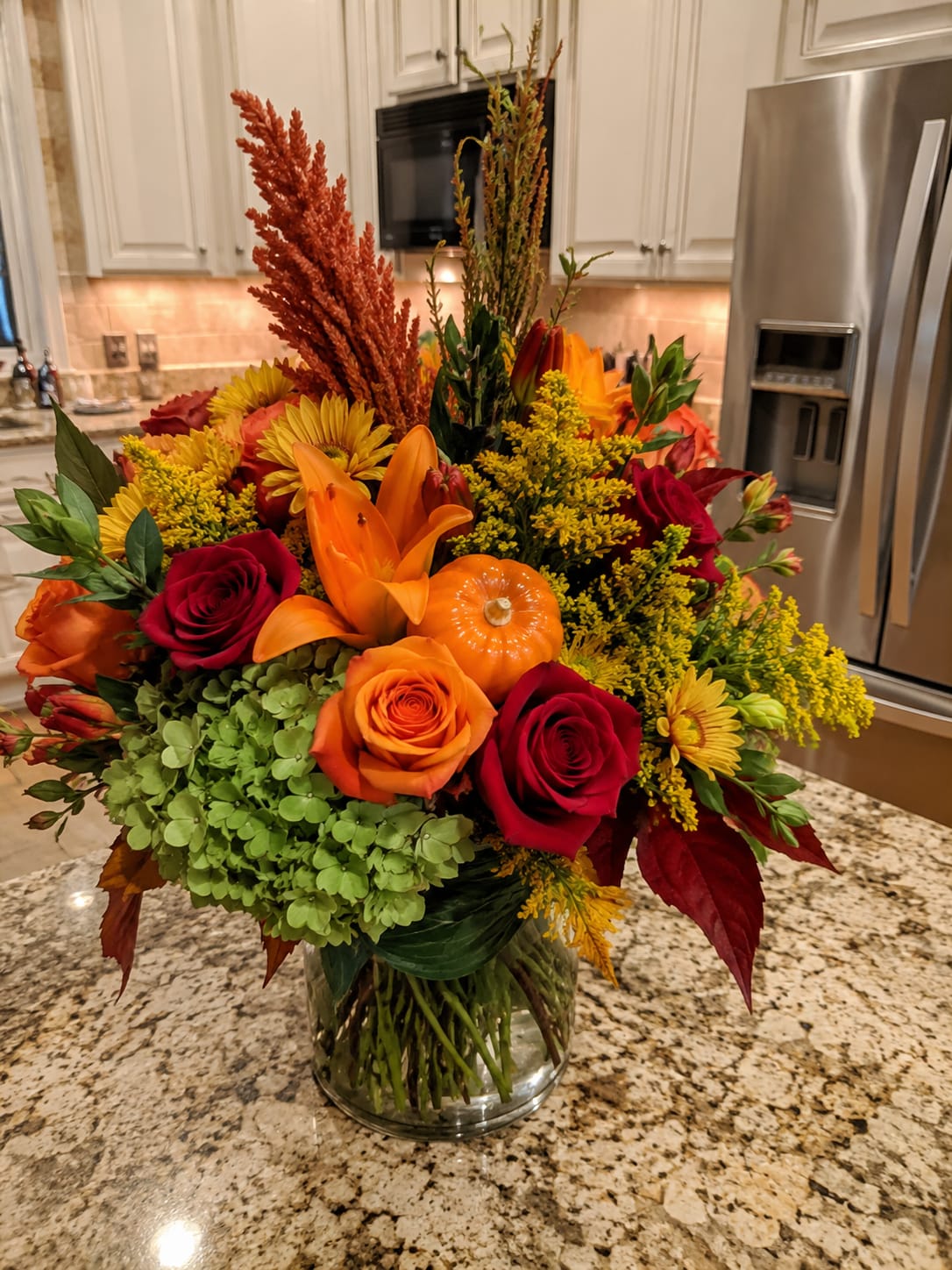Fall Bliss in Palmetto  - A bold burst of autumn color-this fall flower arrangement for Thanksgiving and seasonal gifting pairs orange lilies with red and orange roses to create a warm, celebratory focal point. Yellow chrysanthemums and tall rust-colored accent stems add texture and golden highlights, while a decorative pumpkin tucked into the center makes the autumn theme unmistakable. The palette reads rich and cozy, perfect for a host gift or to surprise someone who loves harvest-season flair.  The design sits in a clear glass vase and features a large cluster of green hydrangea that grounds the arrangement with soft, cool contrast against the vivid oranges, reds, and golds. The composition is full and sculptural, with height from the accent stems and a generous, rounded feel from the roses, lilies, and chrysanthemums. Ideal for a Thanksgiving centerpiece, seasonal mantel display, or to brighten a loved one's home, this arrangement is delivered ready to display; stems are arranged for immediate enjoyment and simple care instructions are included with your order.
