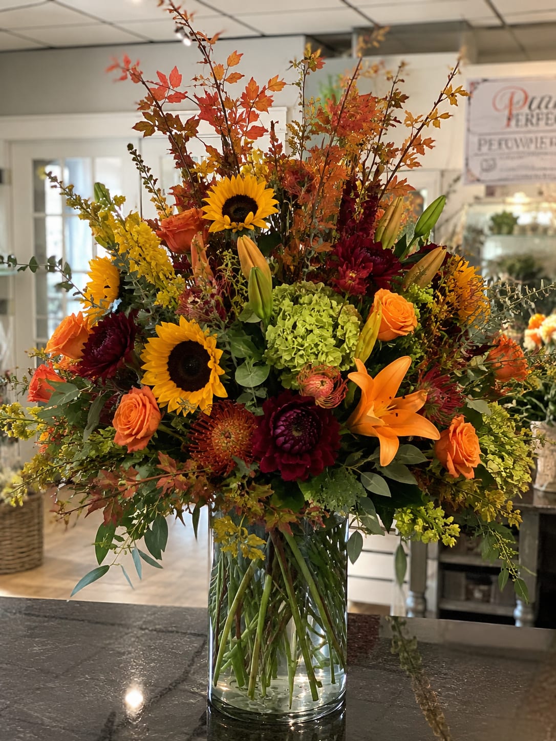 Fall into Fall  - Bright sunflowers and orange lilies set a radiant, harvest-ready tone in this fall flower arrangement, perfect as a Thanksgiving centerpiece or a cheerful birthday bouquet for someone who loves autumn. This flower arrangement for fall blends bold sunflowers, orange roses, and burgundy chrysanthemums to create a warm, seasonal palette that reads festive and heartfelt-the kind of gift that makes the person you're thinking of feel celebrated and cozy.  Arranged in a clear cylindrical glass vase, the design pairs a single cluster of green hydrangea with tall branching stems and mixed textural accents for a full, airy silhouette that feels garden-fresh. The combination of sunflowers, roses, lilies, hydrangeas and chrysanthemums gives visual contrast and depth-bright yellows and oranges against rich red and green-making the display striking from every angle.  Ideal for autumn gatherings, host gifts, or to mark a fall birthday, this abundant arrangement brings harvest-inspired warmth to mantels, dining tables, or entryways. Sized as a large, statement design, it arrives ready to display in the included vase; to keep blooms looking their best, refresh the water every few days and trim stems slightly on delivery.