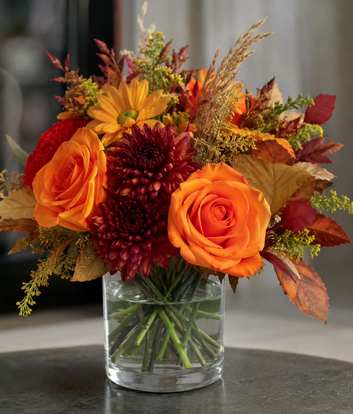 Fall Possies  - Bright orange roses and deep burgundy chrysanthemums set a warm autumn tone in this fall bouquet-perfect as a Thanksgiving centerpiece or a seasonal gift to celebrate harvest-time coziness. The arrangement reads cheerful yet grounded, the kind of floral surprise that will make the person you're thinking of feel noticed and comforted by rich, autumnal color.  Mixed yellow accents, red foliage, and dried grass-like stems add texture and height, creating a full, layered look in a clear cylindrical glass vase. Colors include orange, red, yellow and mixed tones; flowers are roses and chrysanthemums. Sized to sit comfortably on a dining table or entry console, it arrives ready to display and brings long-lasting seasonal charm to any room.