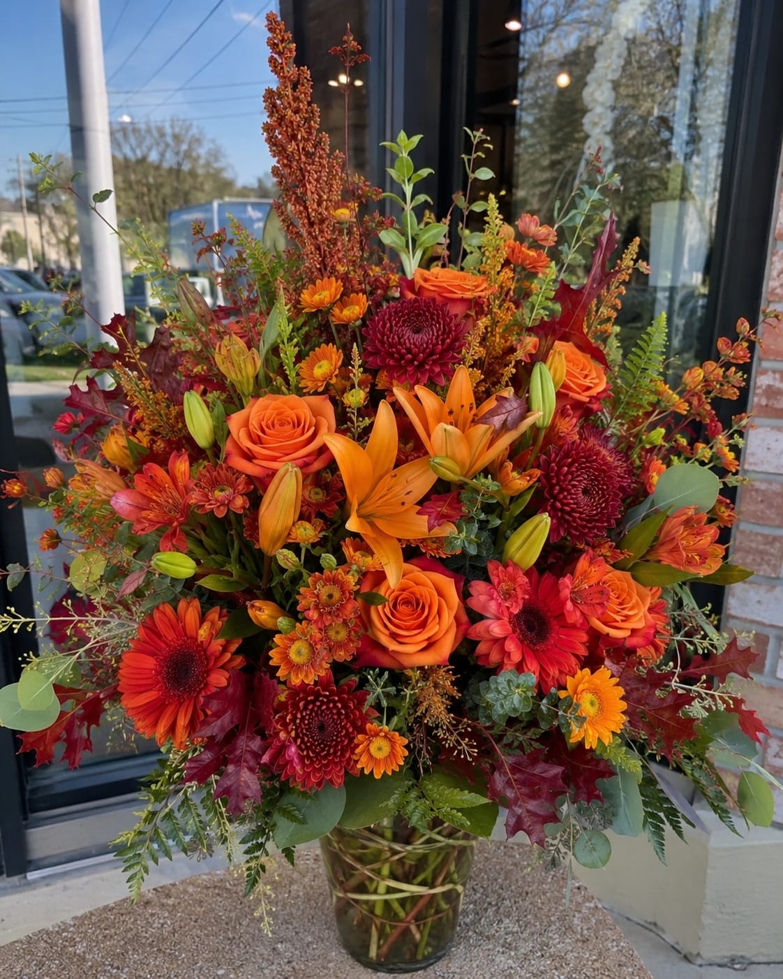 Fall Romance in Brandon  - A dramatic autumn centerpiece, this large fall flower arrangement is a bold choice for Thanksgiving, a seasonal celebration, or an autumn birthday. The upright vase design centers on orange roses and orange lilies with deep red chrysanthemums to create a warm, romantic feel-perfect for the person you want to make feel cherished this season.  Packed tightly in a clear glass vase, the arrangement mixes gerbera daisies, alstroemeria spray blooms, and chrysanthemums to build a dense, layered texture of burgundy, rust, gold, orange, and touches of green. The varied shapes-from the round roses and daisies to the sculpted lilies-add rich visual contrast and a full, abundant presence that reads like a harvest table focal point.  Made to impress on arrival, this arrangement arrives ready to display as a centerpiece or gift. It's sized as a large upright design; keep it in fresh water and out of direct heat to extend vase life so the recipient can enjoy its autumn scent and color for days.