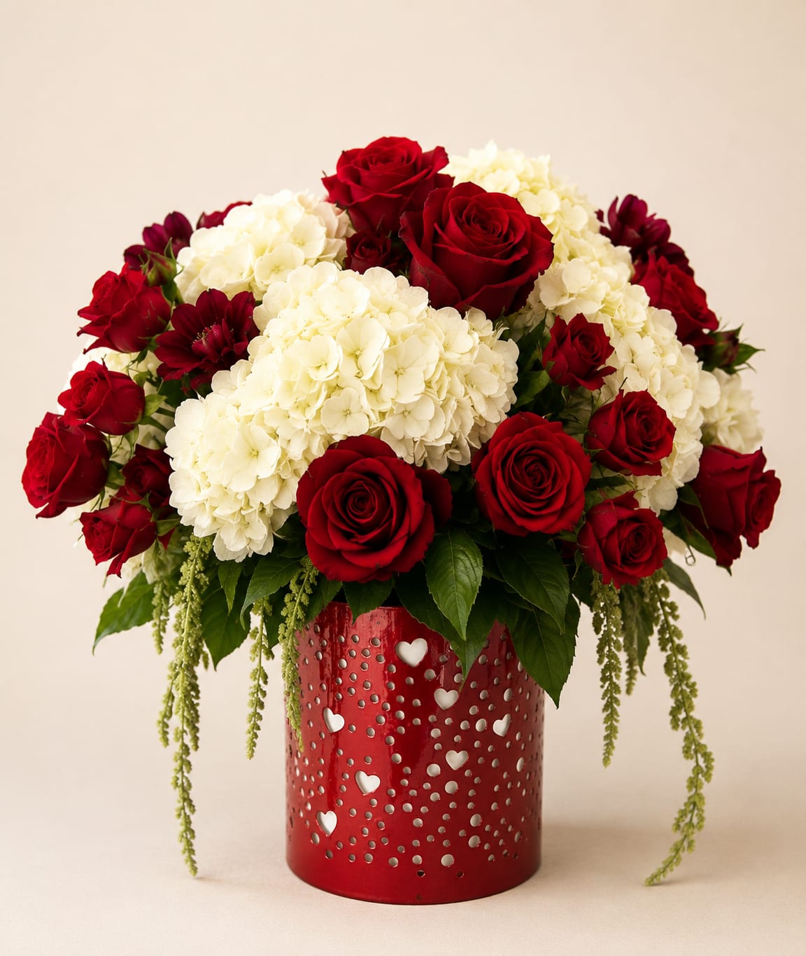 Loves In The Air - Deep red roses paired with voluminous white hydrangeas create an unmistakably romantic statement in this flower arrangement-perfect as a birthday bouquet for a sweetheart or an anniversary roses gift that says "I love you." The saturated crimson and creamy white contrast draws the eye and feels polished yet heartfelt, making the recipient feel truly cherished the moment they see it.  The arrangement features lush clusters of white hydrangeas surrounding rich red spray roses for a full, rounded silhouette. Fresh greenery adds texture and a natural frame, and the blooms are presented in a glossy red metal vase with heart-shaped cutouts that reinforce the love-themed design. The vase's sturdy cylinder shape gives the display a modern, celebratory look and makes it easy to place on a table or mantel.  Ideal for romantic occasions, Valentine's Day, anniversaries, or simply to say you're thinking of them, this arrangement delivers an elegant, emotional impact. Size and freshness make it suitable for gifting to the person you want to feel adored.
