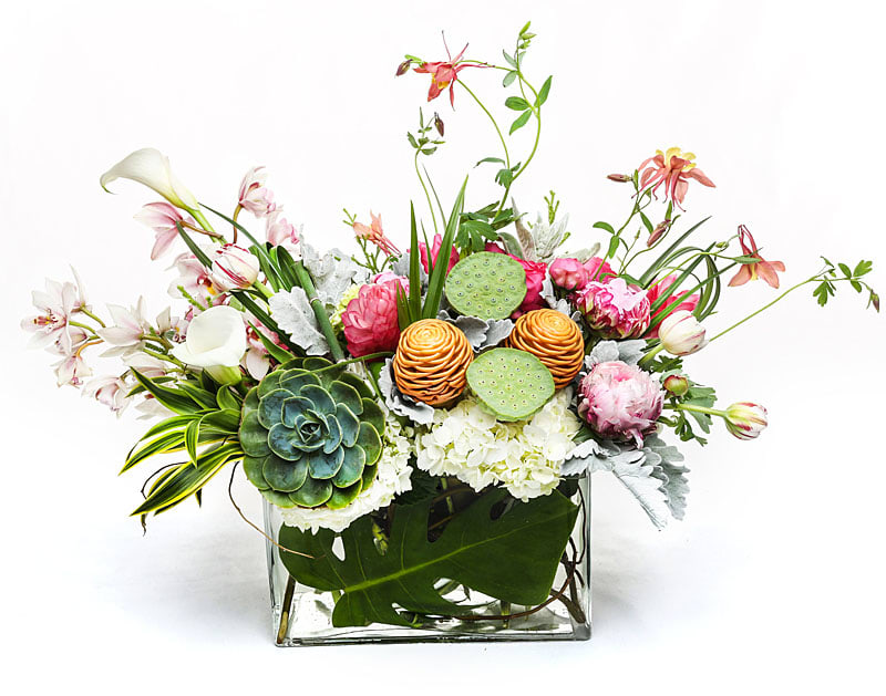 Mixed floral arrangement in a clear square vase with lotus pods and succulents