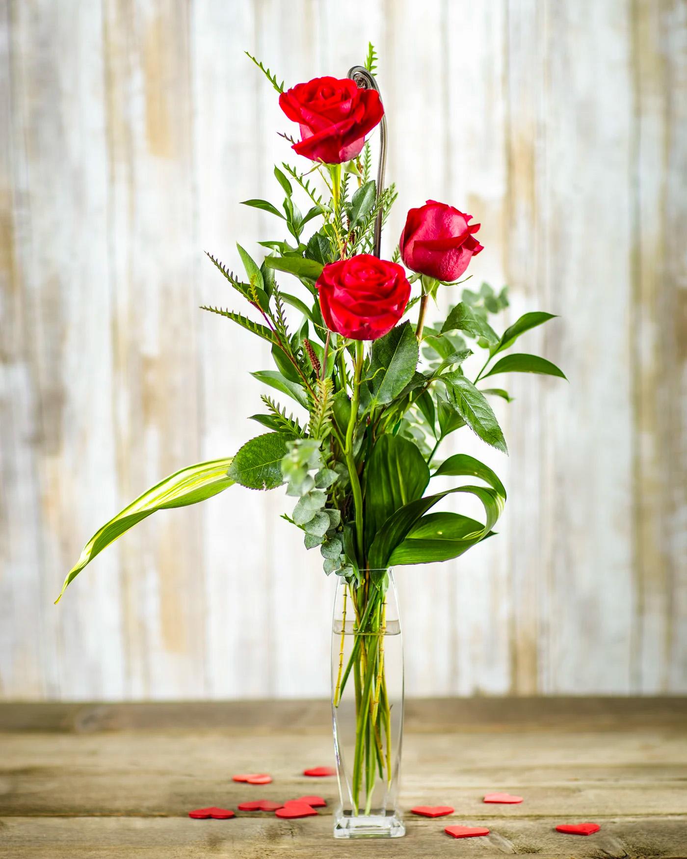 Red roses in a tall clear vase with scattered heart confetti