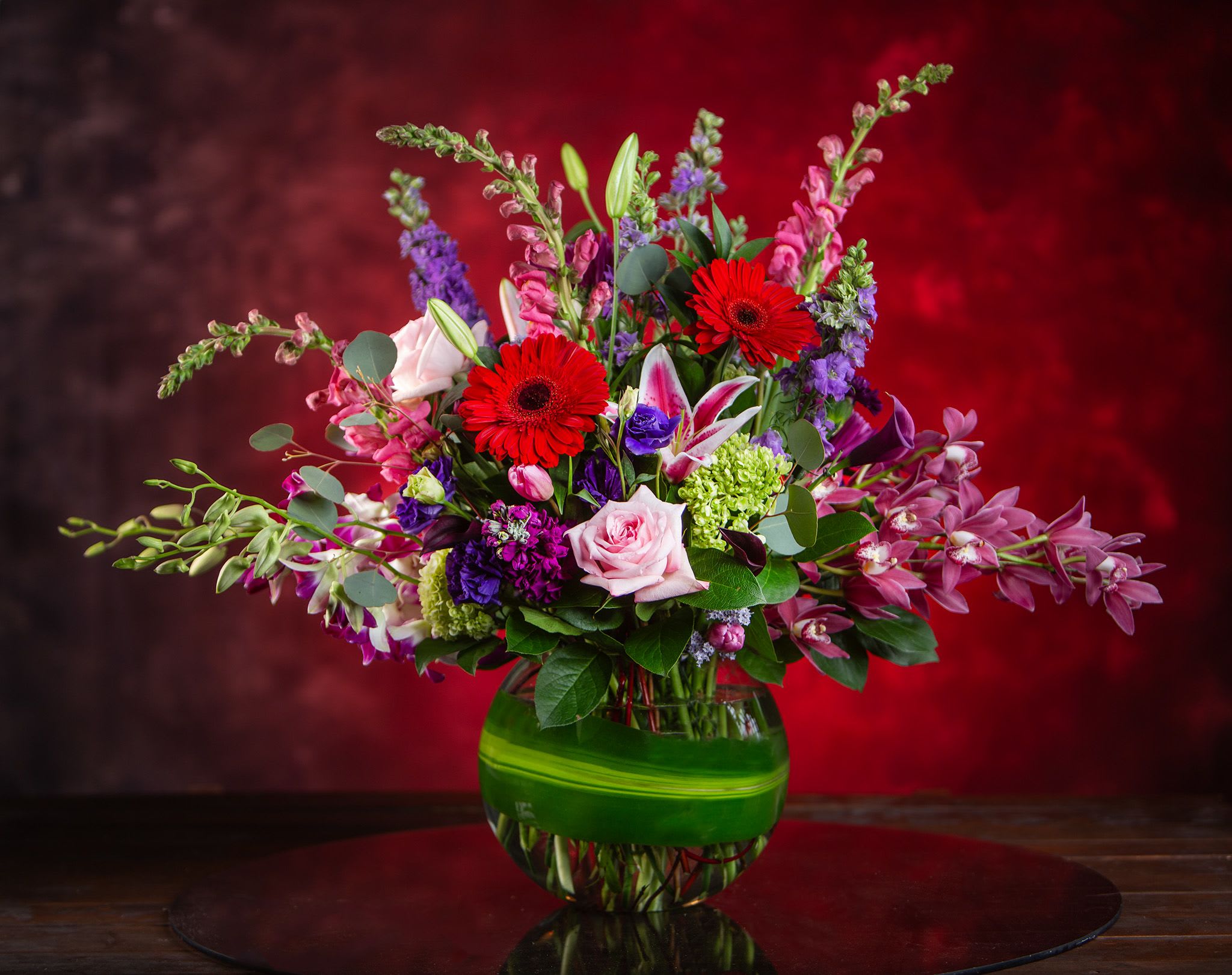 Mixed bouquet with red gerbera daisies, pink roses, and purple blooms in a green glass vase