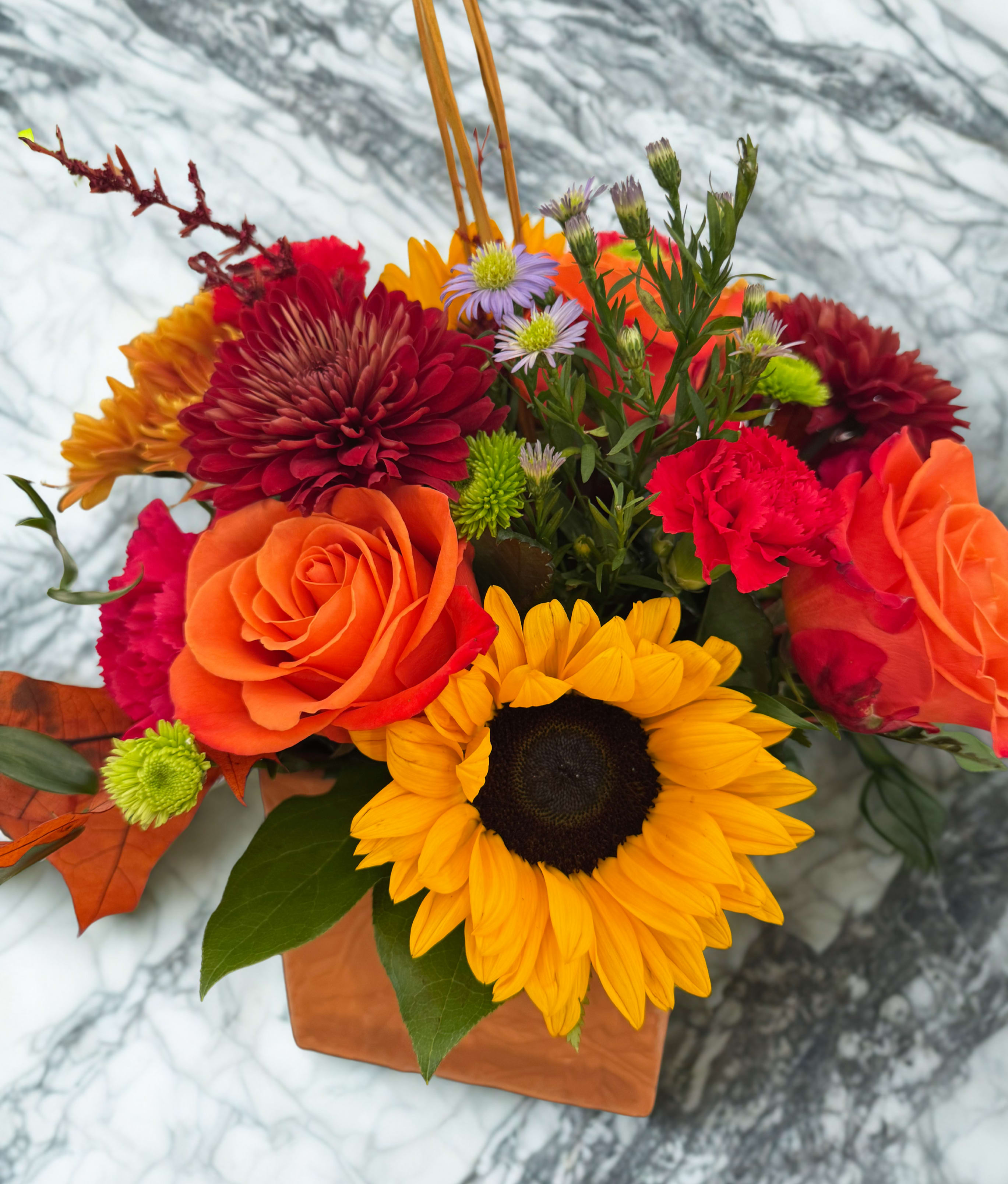 Our Version of the FTD Outdoors Bouquet - Bring the beauty of the season indoors with this warm and welcoming autumn arrangement. Vibrant sunflowers, glowing orange roses, and rich burgundy mums come together to create a stunning display inspired by the colors of the outdoors. Each bouquet is artfully designed in a charming terracotta cube embossed with a leafy motif, adding rustic elegance to any space. Deluxe size: Approximately 12" H x 14" W.  $55.00, $65.00 and $70.00