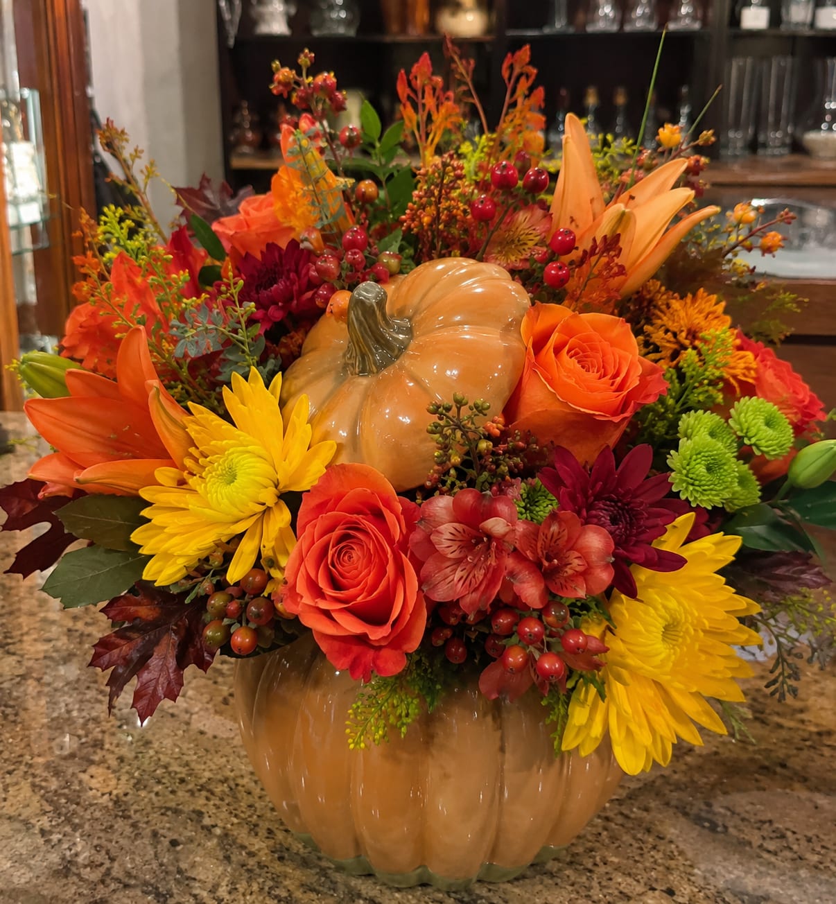 Harvest Pumpkin for Ruskin - Glossy pumpkin-shaped ceramic vase at the center gives this fall flower arrangement its unmistakable harvest-table charm - a seasonal floral centerpiece for Thanksgiving, autumn decorating, or a warm fall gift. The low, rounded silhouette and packed, abundant design create a welcoming, homey feel that will make the person you're thinking of feel celebrated and cozy.  A dense mix of orange roses, orange lilies, yellow and burgundy chrysanthemums, and red alstroemeria is layered with berry accents and fall foliage for rich texture and depth. Warm oranges, golden yellows, deep reds, and autumn greens read beautifully together against the pumpkin vase, creating a festive, seasonal palette that looks lovely on a dining table or mantel.  Perfect for saying thanks, marking a fall birthday, or adding seasonal cheer to someone's home, this arrangement arrives ready to display in its ceramic pumpkin - decorative and substantial rather than formal. For best enjoyment, keep flowers in fresh water and out of direct heat to prolong bloom life; available in a single low, rounded size ideal for centerpieces.