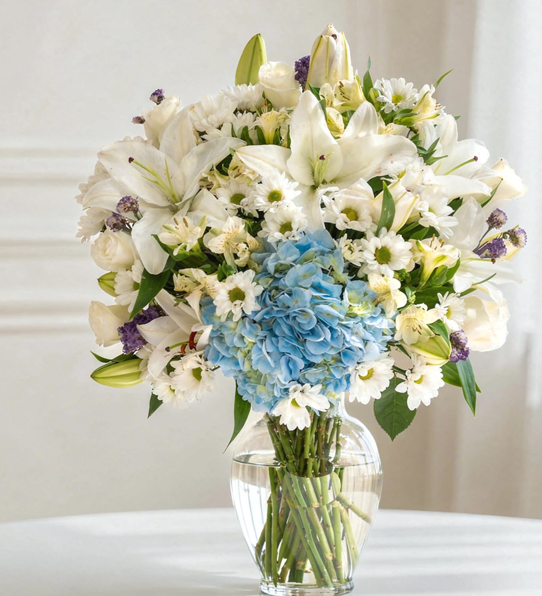 A serene and elegant vase  - A serene and elegant vase arrangement featuring 3 stems of alstroemeria, 4 stems of white chrysanthemums, 3 graceful white roses, 2 stems of white lilies, vibrant blue hydrangea, and accents of purple statice. Designed in a clear glass vase, this arrangement blends soft whites with gentle pops of blue and purple, creating a calming yet refined floral display—perfect for modern interiors and thoughtful gifting.  