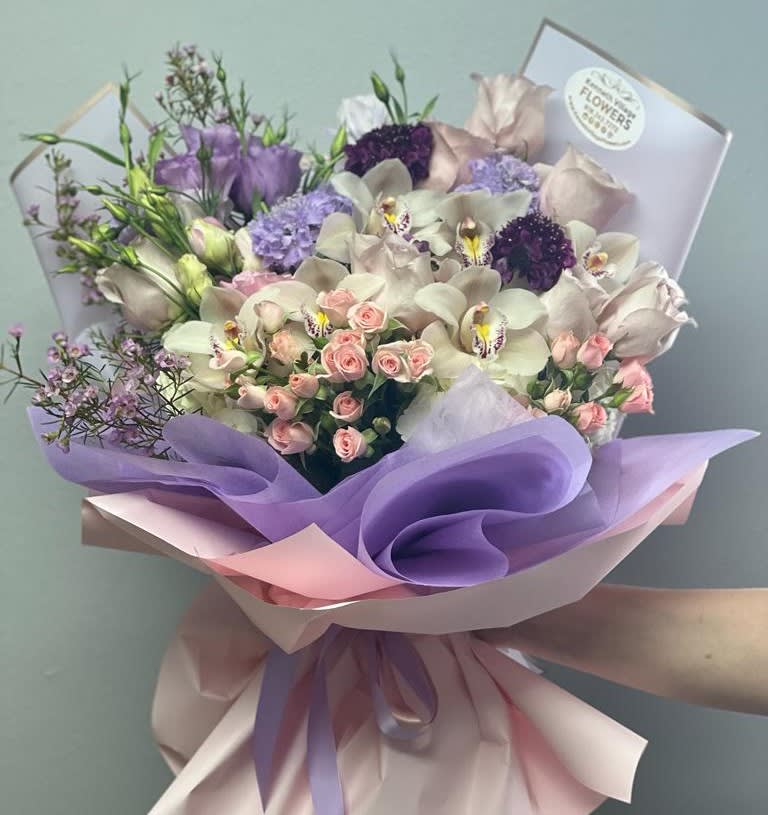 Bouquet of pastel orchids, roses, and purple blooms wrapped in lavender paper