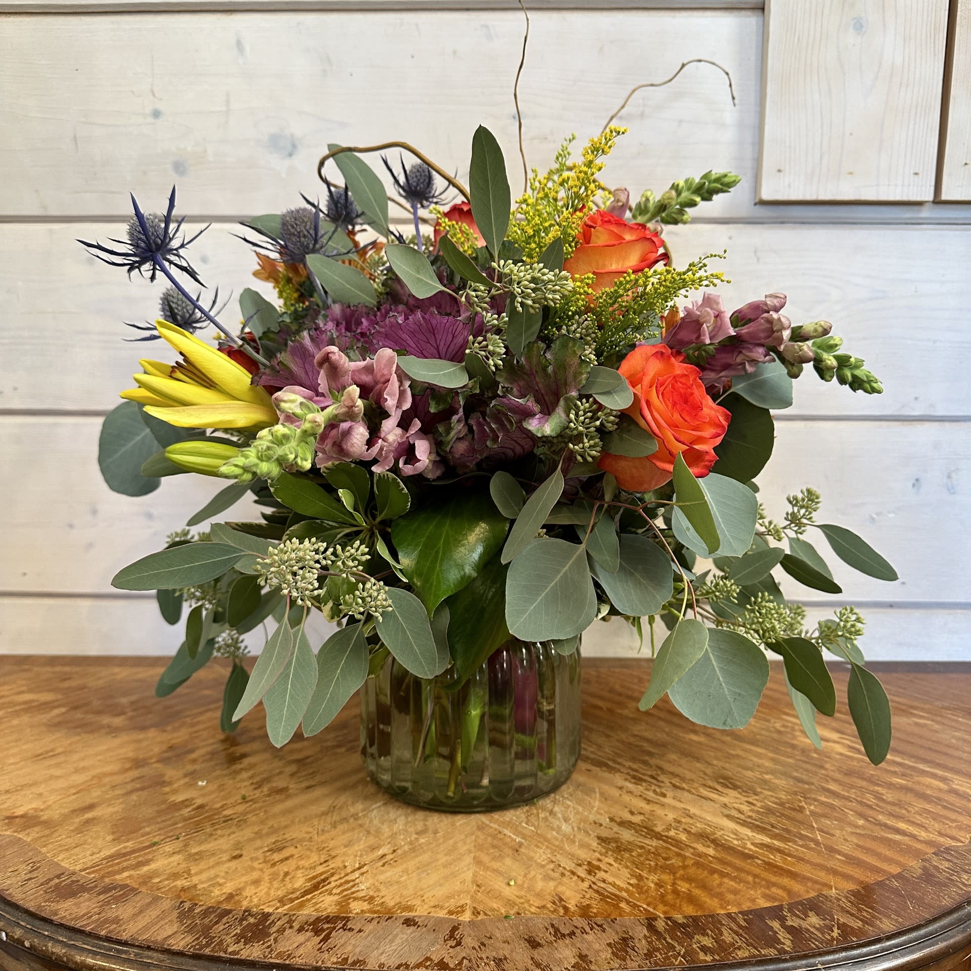 Mixed bouquet in a glass vase with orange roses, yellow blooms, and purple flowers