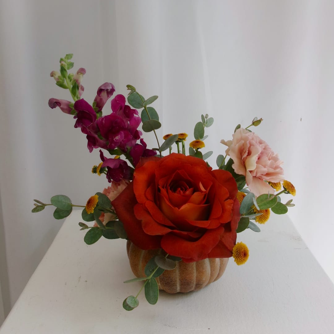 Small floral arrangement with a red rose, purple snapdragons, and pale pink carnations in a pumpkin vase