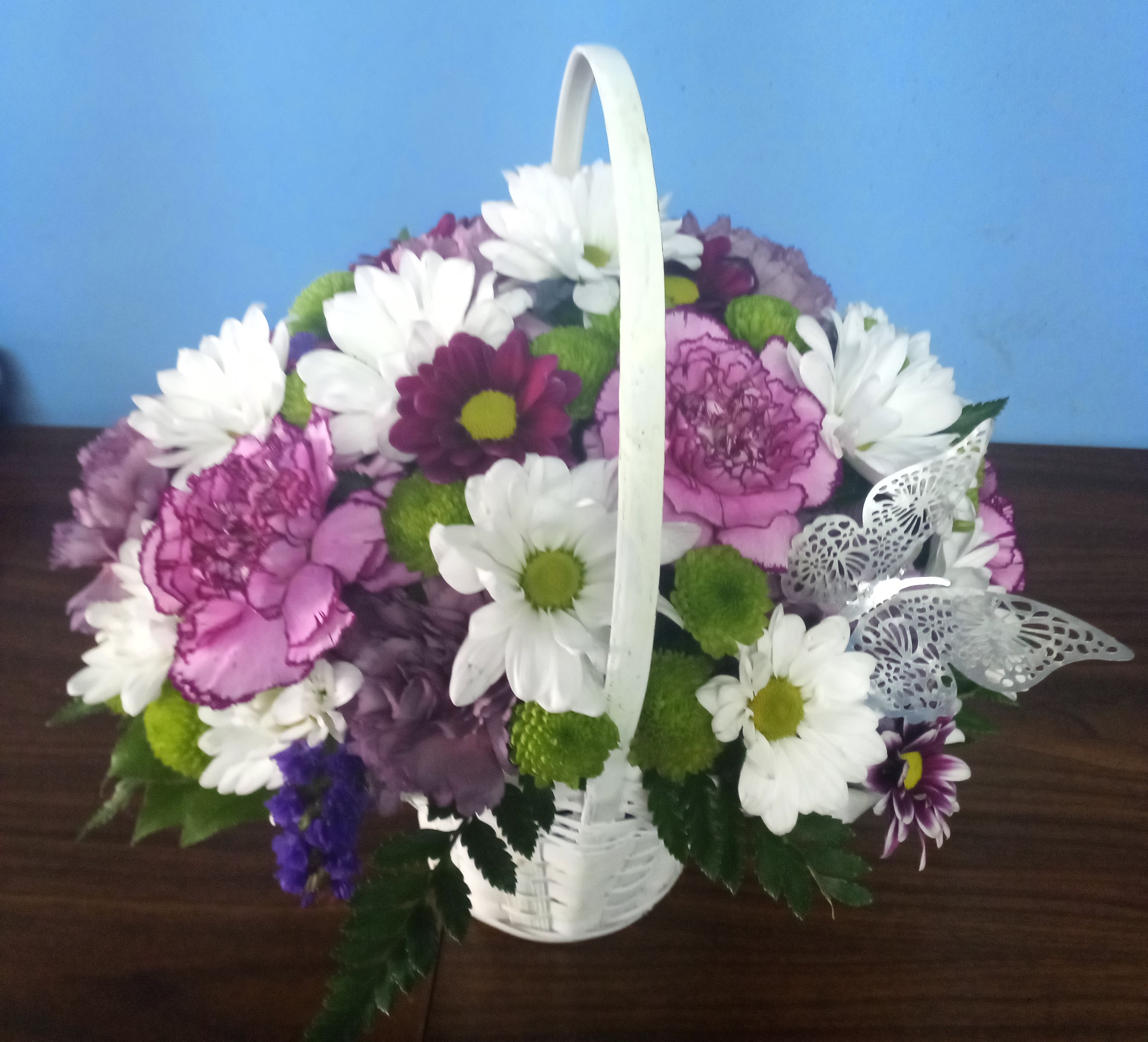 Blooming Bounty - This pretty basket is sure to cheer up anyone's day. This white-wash handled basket is filled with green button pompons, white daisy pompons, hot pink mini carnations and purple carnations. A lavender ribbon bow completes your best wishes.