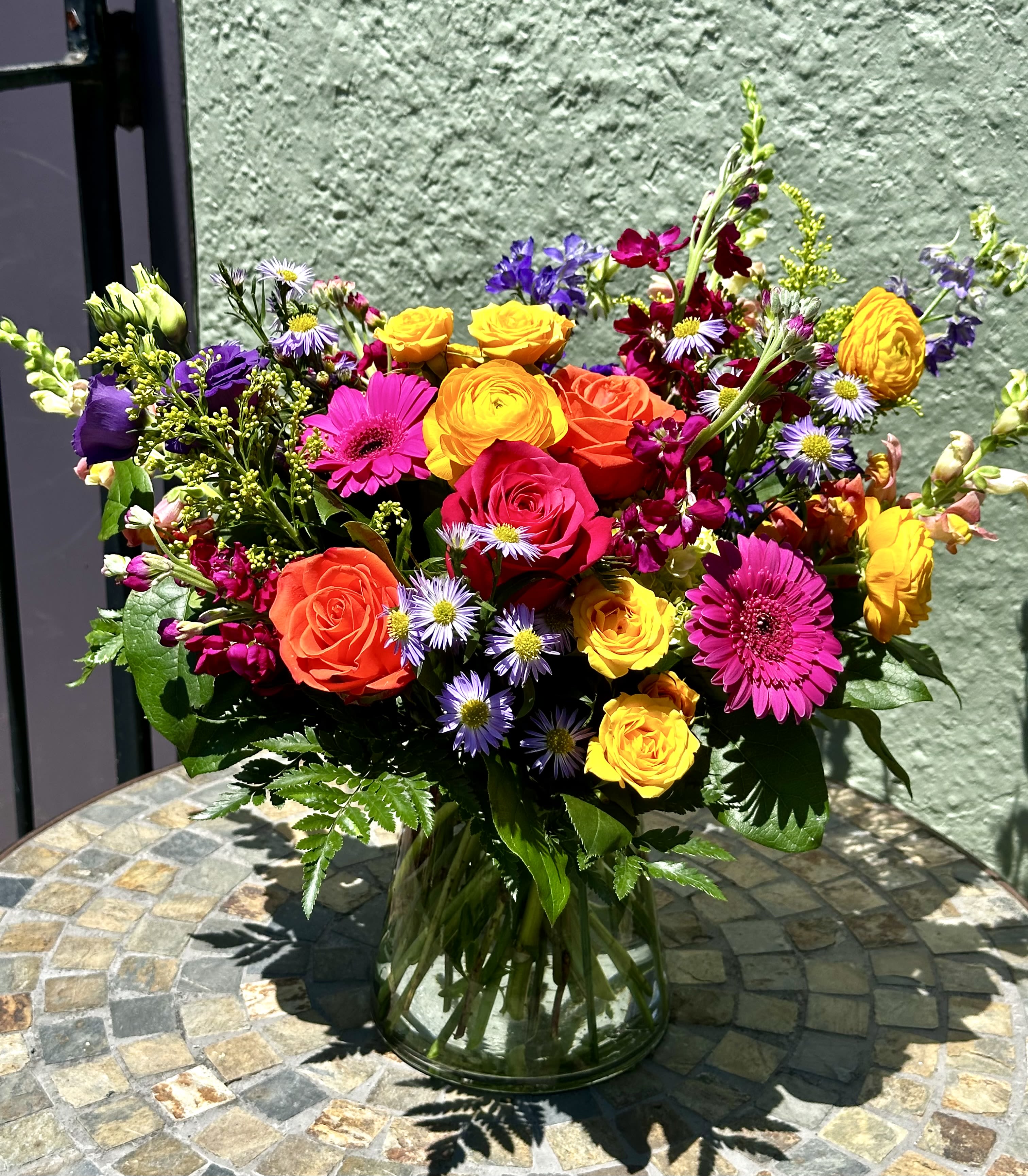 Rainbow Connection - A joyful celebration of summer, Rainbow Connection features a diverse array of vibrant blooms including roses, gerbera daisies, and ranunculus. Designed with full and layered texture, it captures the beauty of a bright rainbow bursting through the sky.