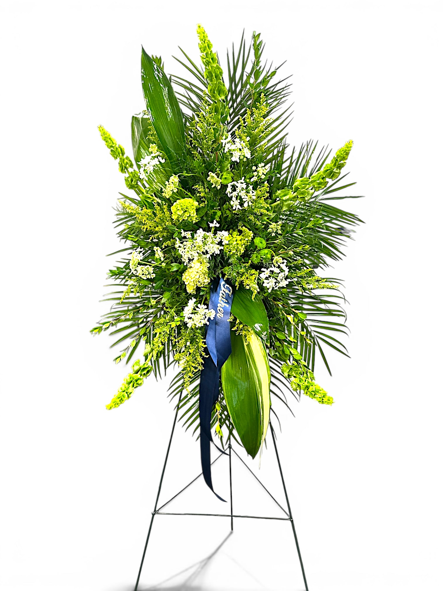 Green Fields Easel - A serene tribute, "Green Fields" features lush greenery and delicate white blooms arranged on a standing easel. Inspired by peaceful open fields, this arrangement offers a sense of tranquility and natural beauty, honoring a loved one with grace and elegance. Perfect for memorial services, it conveys heartfelt remembrance and comfort.