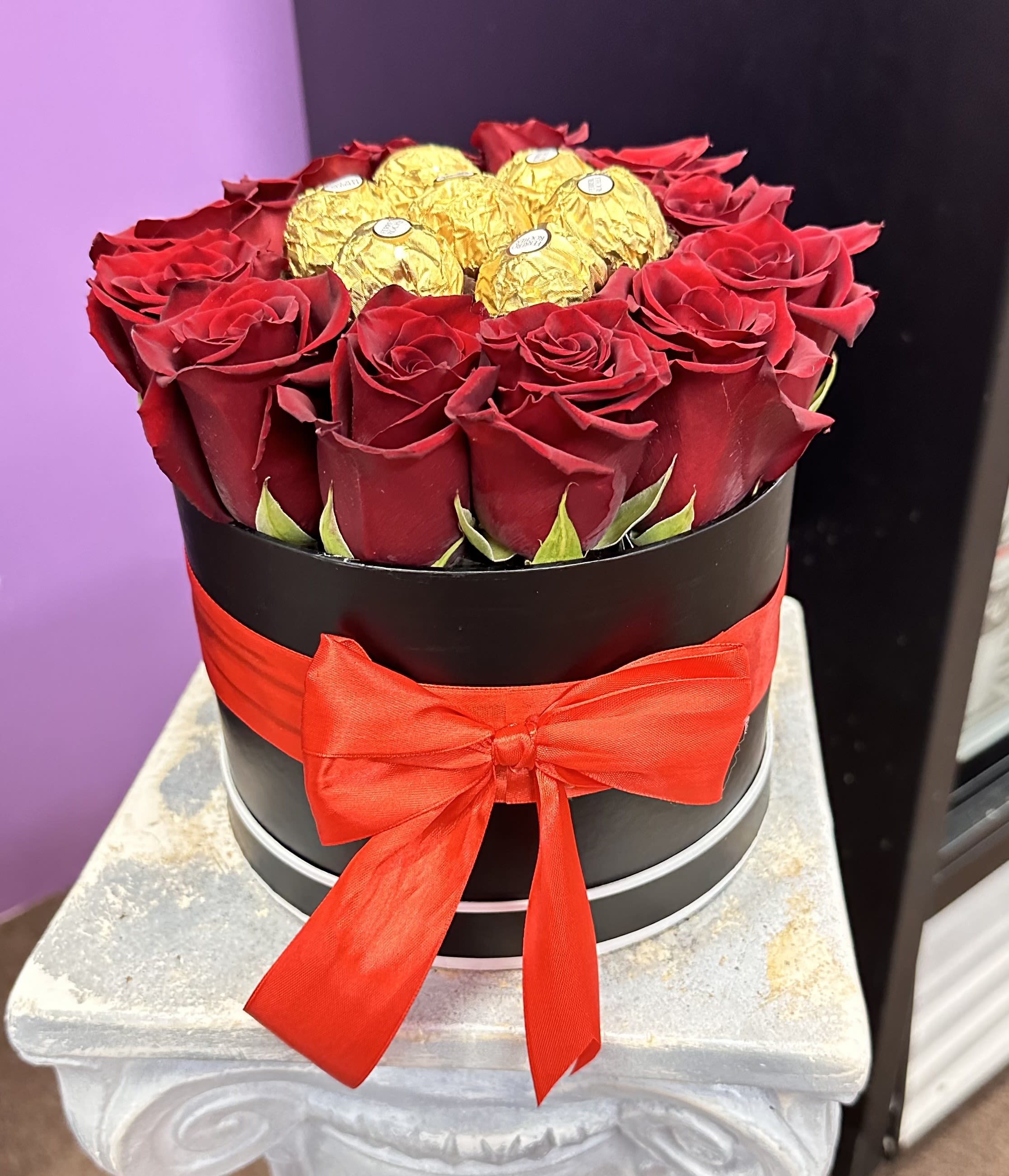 Red roses arranged in a black round box with gold-wrapped chocolates and a red ribbon.