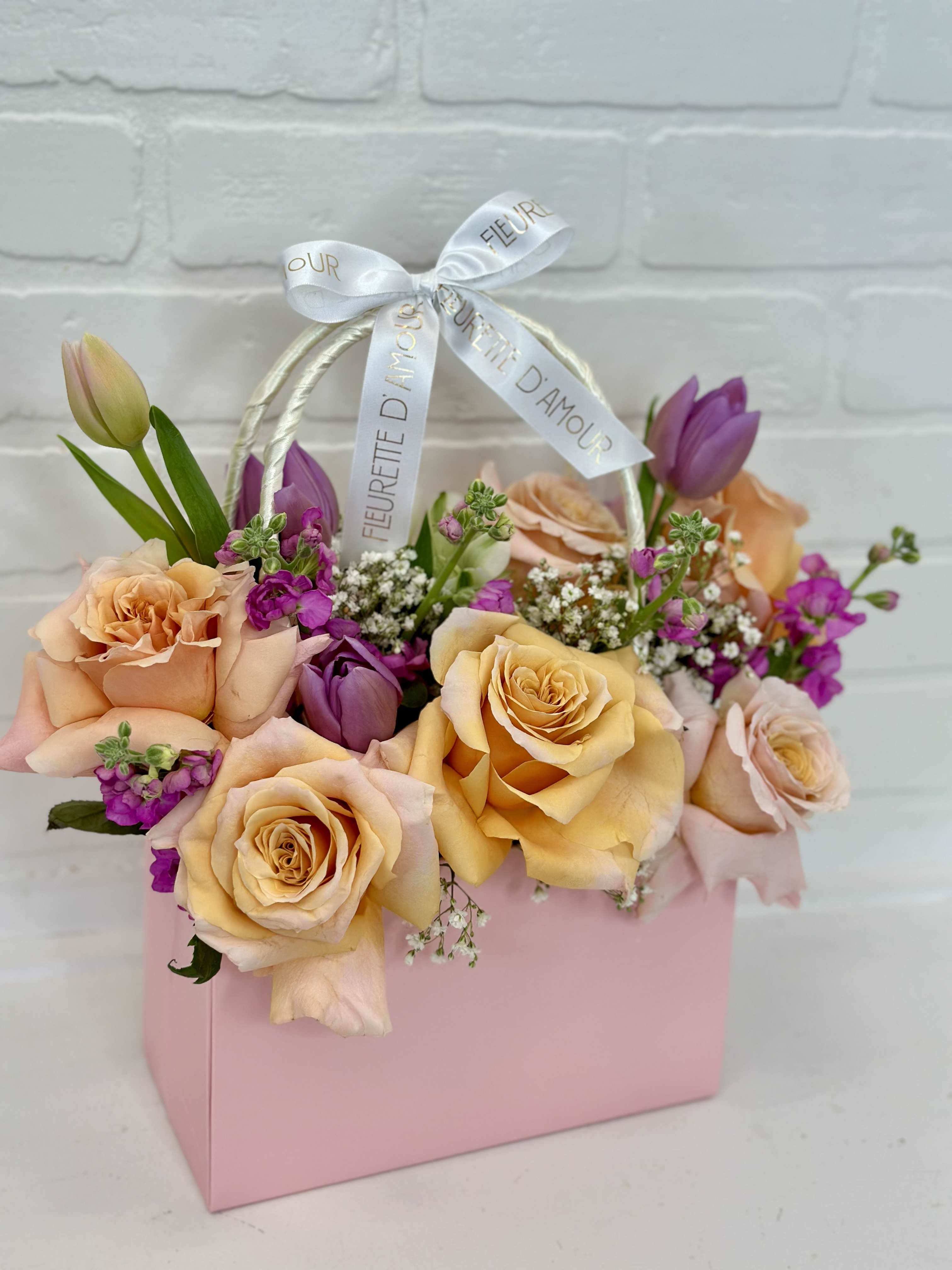 Peach and purple roses arranged in a pink gift box with a ribbon handle.