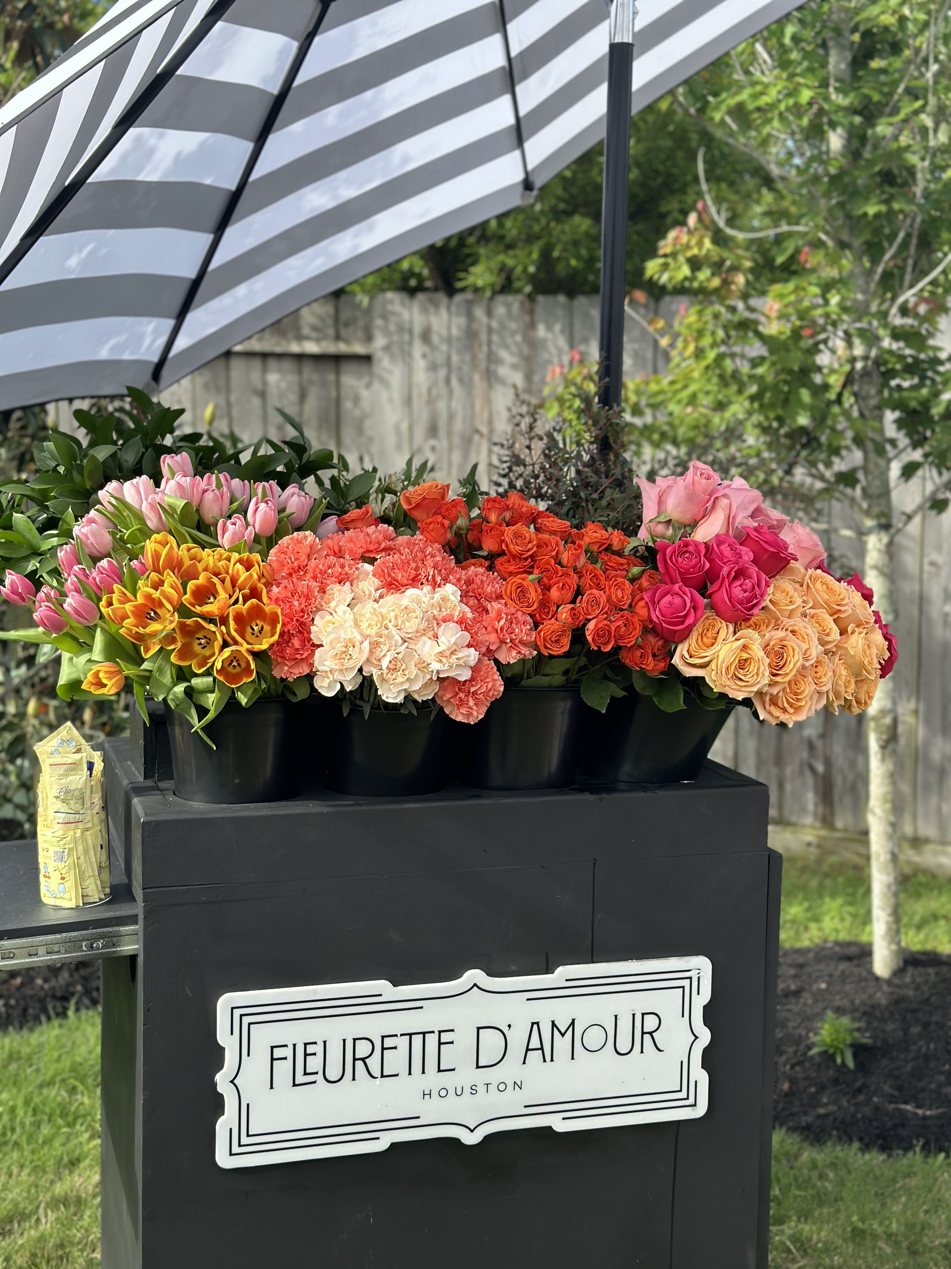 Colorful flower buckets displayed under a striped umbrella outdoors