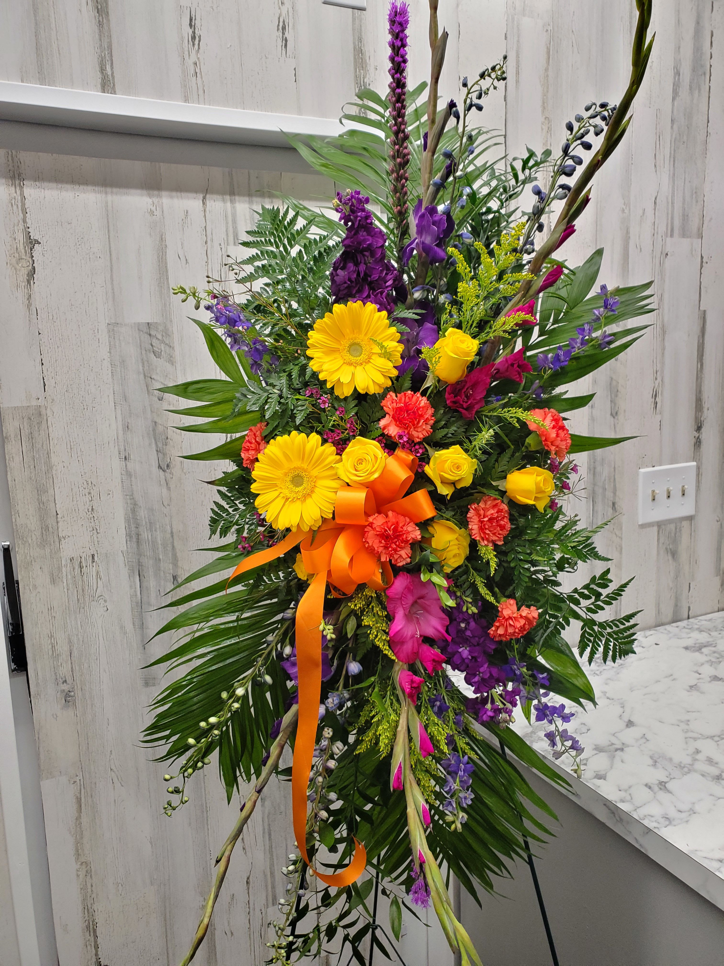 Celeste - One standing spray arrives with orange gladioli and roses, yellow gerberas, hot pink asters and foliage accents on an easel.