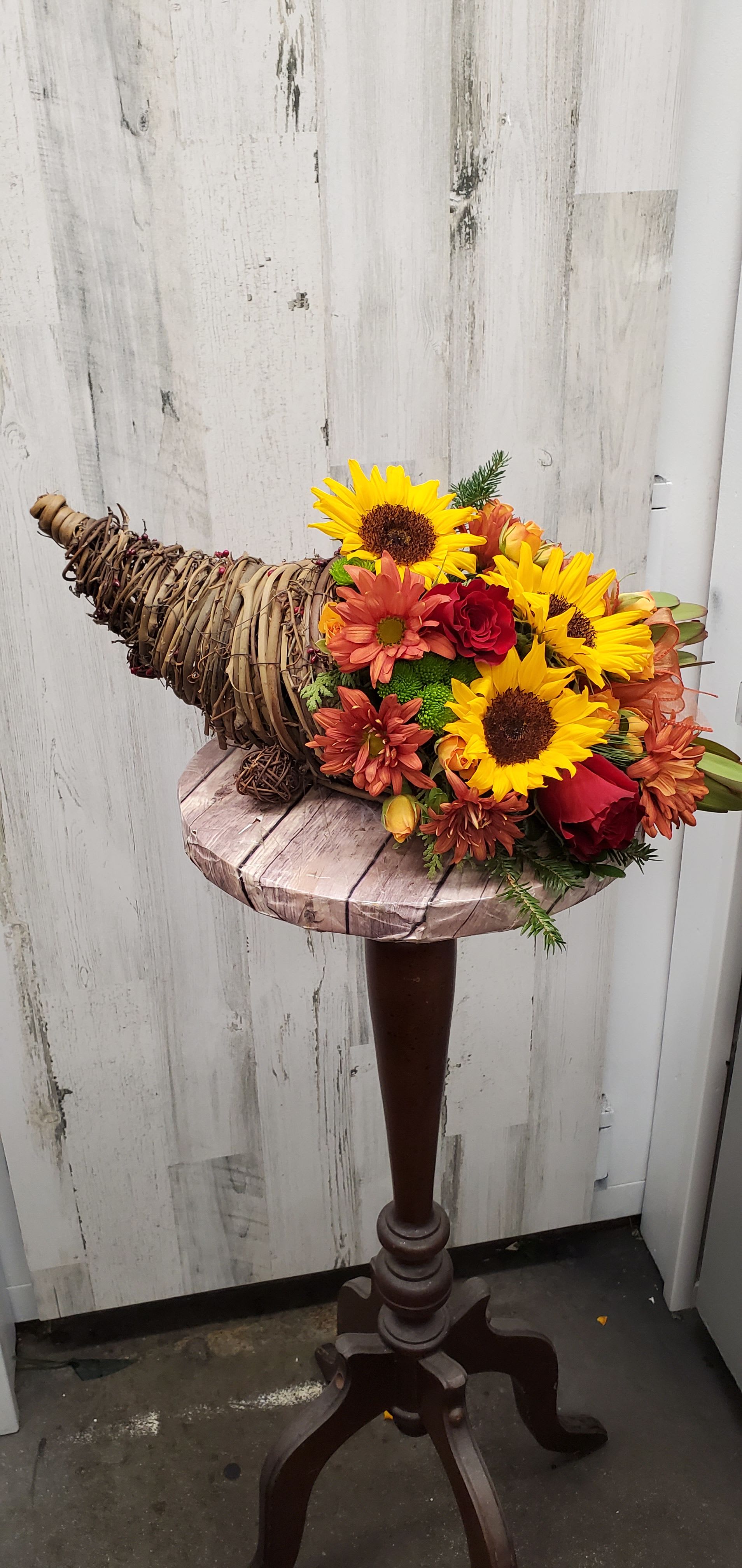 Classic Cornucopia - Along with joy, this cornucopia carries an abundance of beautiful fall flowers and foliage. A stunning centerpiece or inviting entryway display, this beauty will be at home anywhere in the house.  Dazzling sunflowers, light orange roses and spray roses, red daisy spray chrysanthemums, yellow cushion spray chrysanthemums, eucalyptus, magnolia leaves, cinnamon sticks and wheat are perfectly arranged in a wicker cornucopia. Now you know why cornucopia translates to "horn of plenty."  Approximately 19" W x 15" H  Orientation: One-Sided  As Shown : T168-3A Deluxe : T168-3B Premium : T168-3C