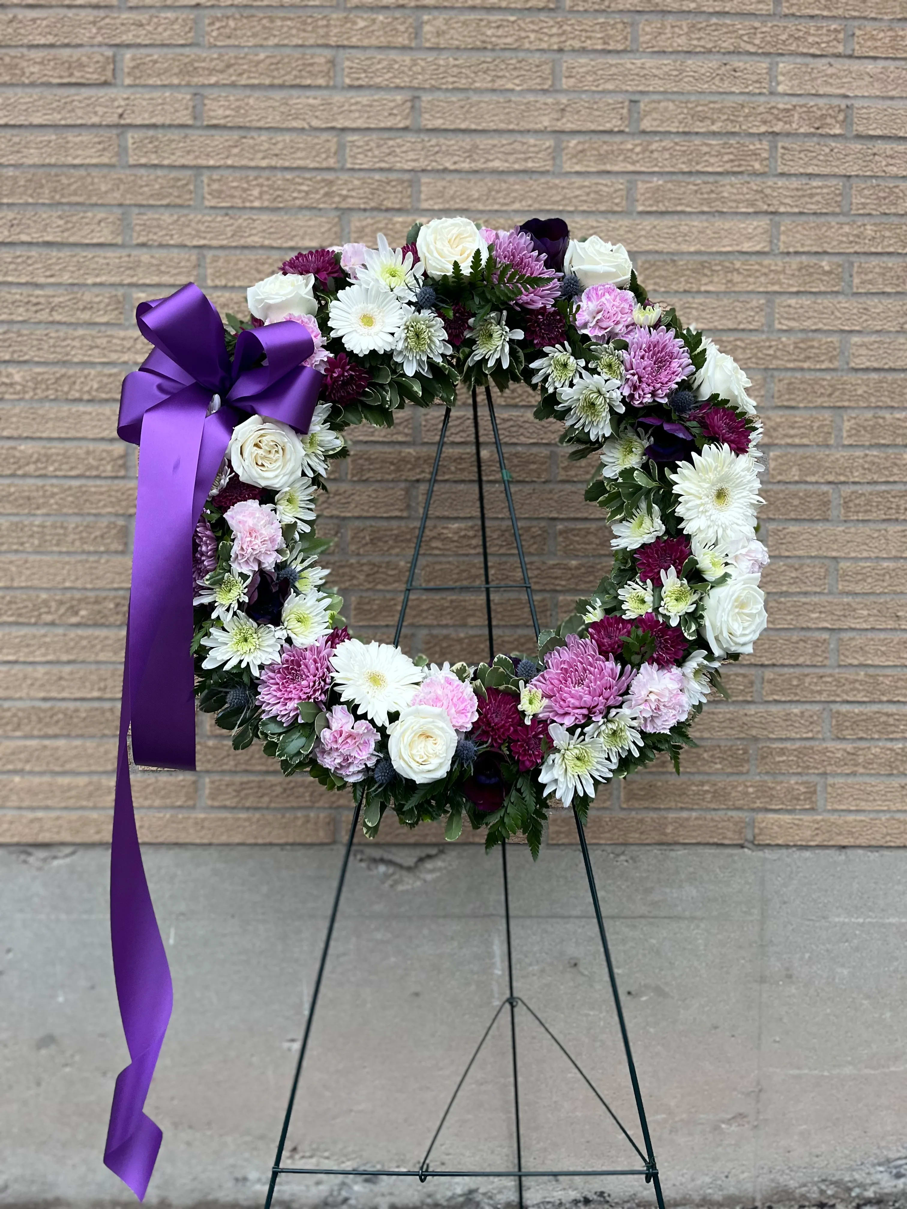 Circular floral wreath with white, pink, and purple blooms on a stand with a purple ribbon