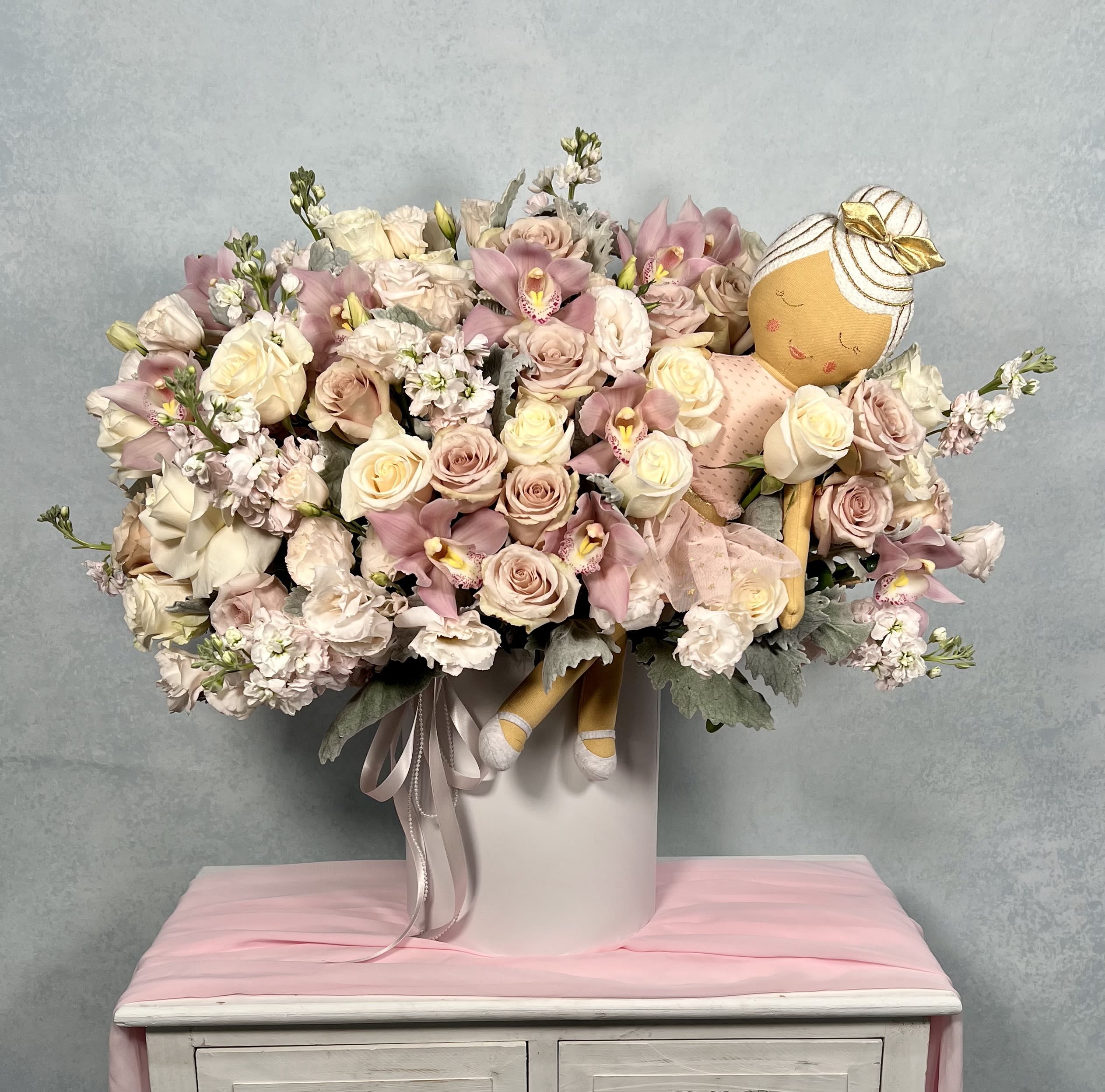 Large pastel pink and cream flower arrangement in a white hatbox with a plush ballerina doll nestled among the blooms