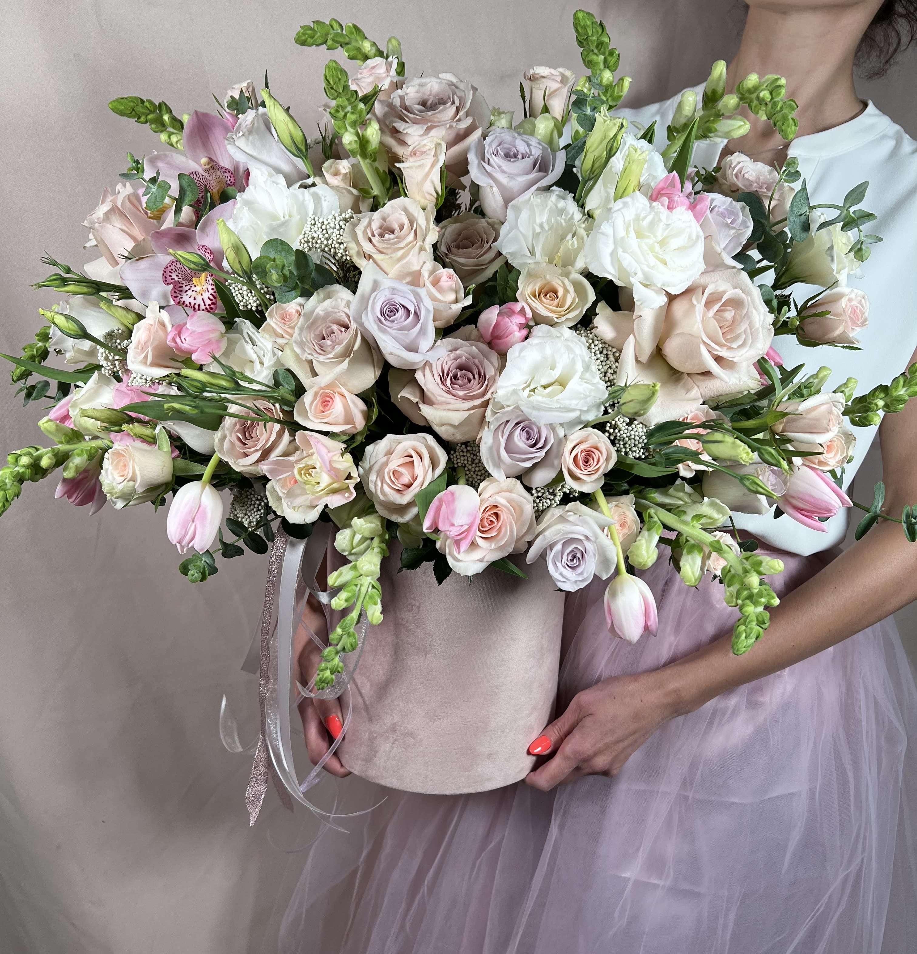 Large pastel arrangement of roses, tulips, and orchids in a blush hatbox held by a person