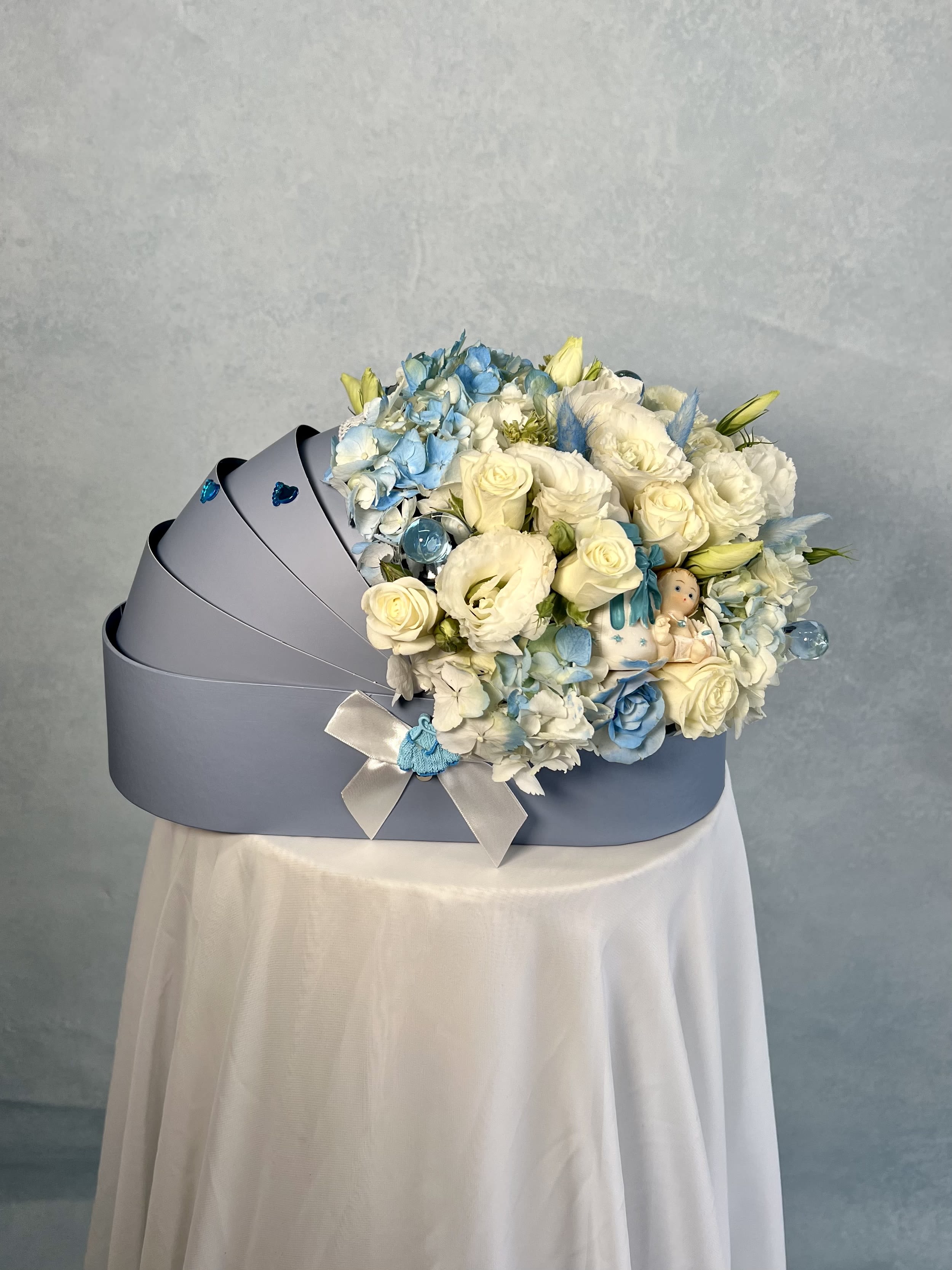 Light blue cradle-shaped box filled with white and blue flowers and a small baby figurine.
