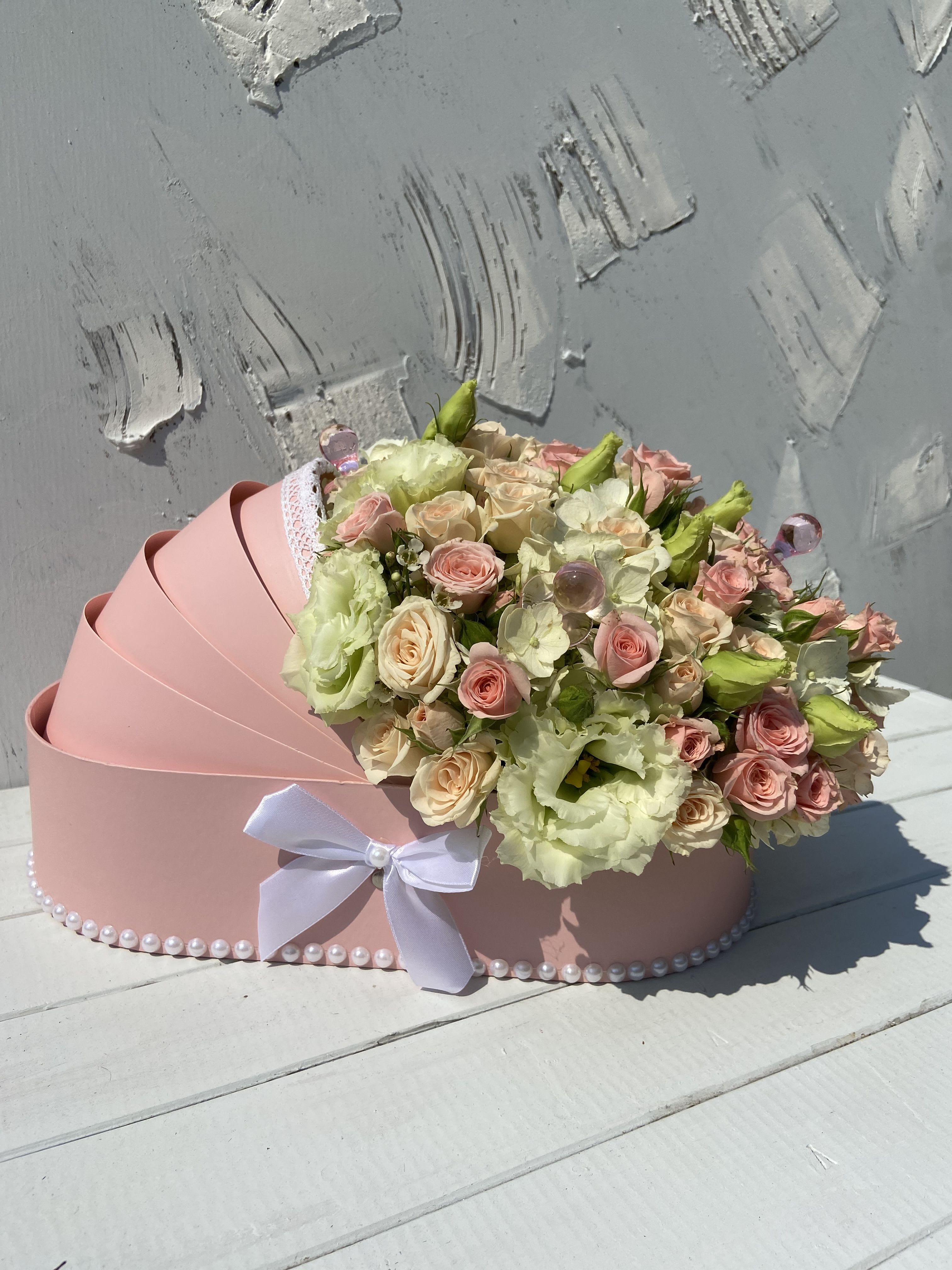 Pink carriage-shaped box filled with cream and blush roses and lisianthus, trimmed with a white bow and pearls