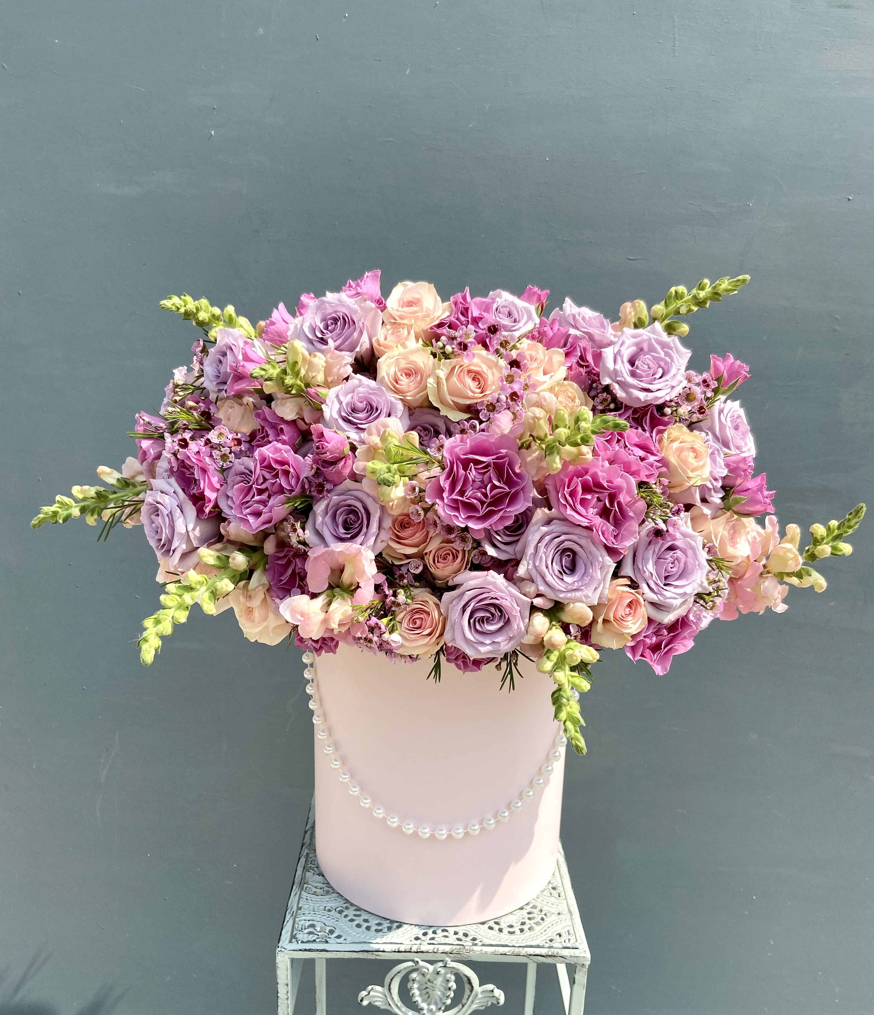 Lavender and pink rose arrangement in a pink hat box with pearls on a decorative metal stand.