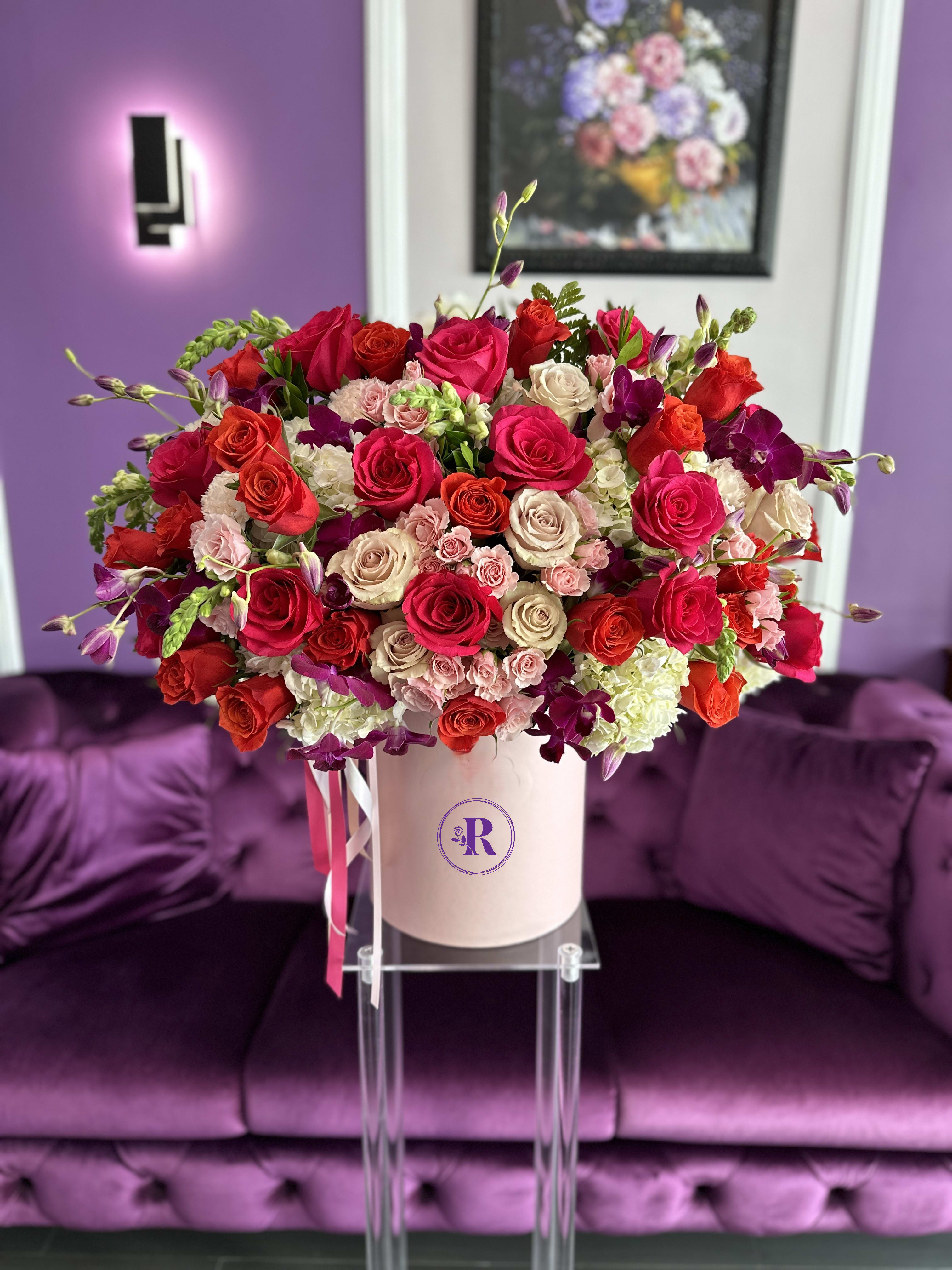 Large hatbox arrangement of red, hot pink, and cream roses with orchids in a blush pink box.