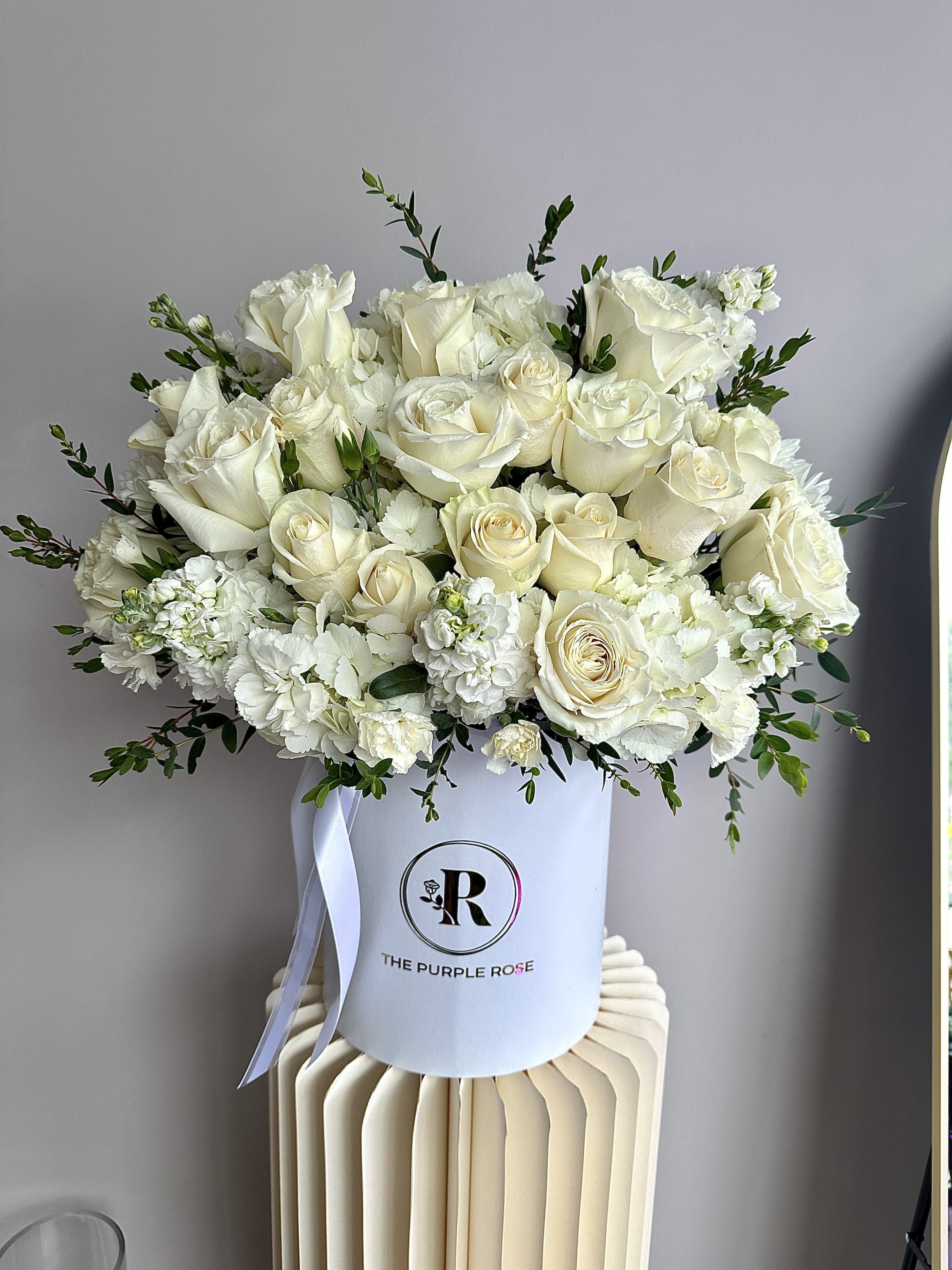 Round hatbox arrangement of ivory roses and white flowers with a ribbon in a white container