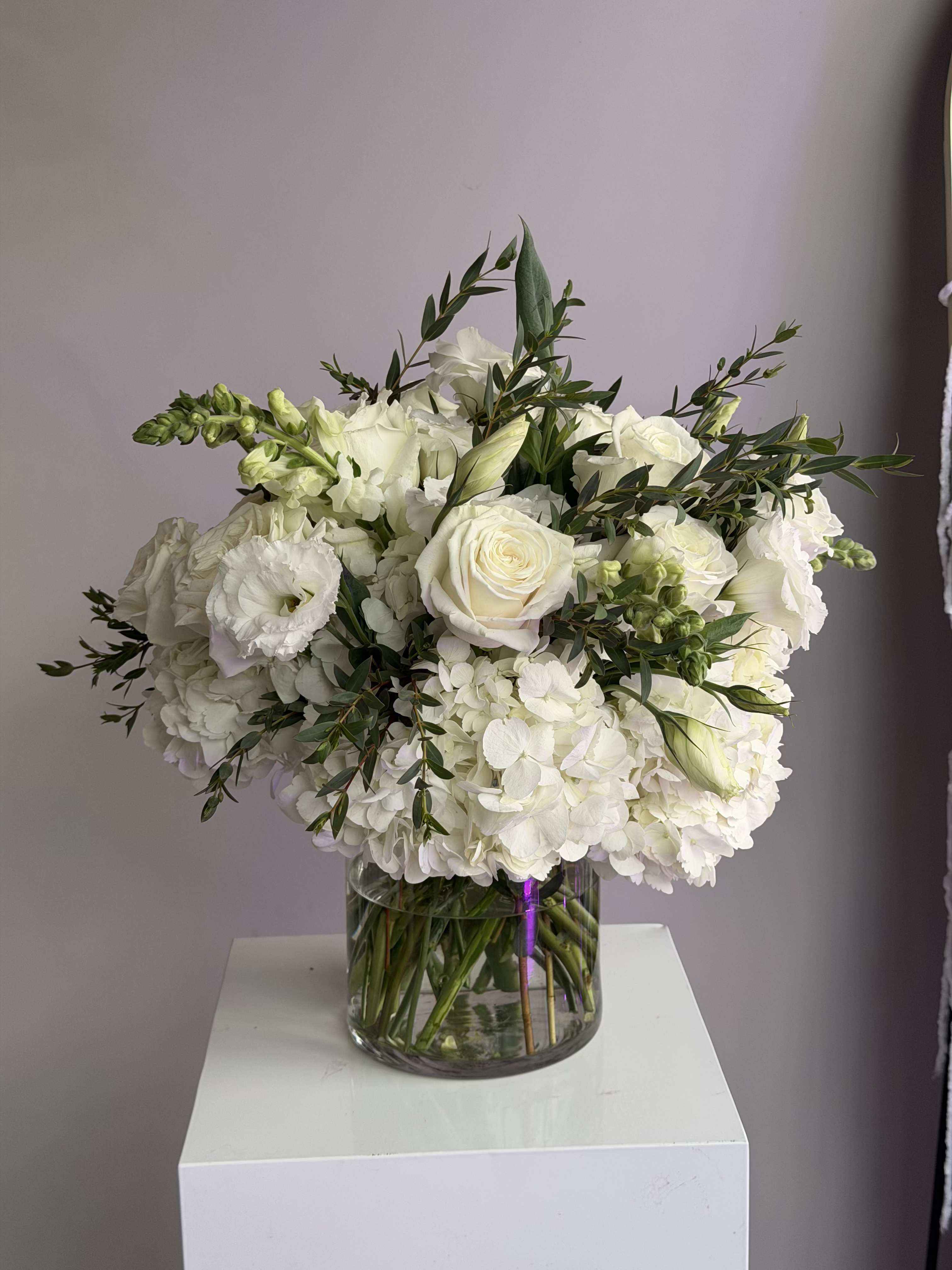 Round arrangement of white roses, hydrangeas, and other white blooms in a clear glass vase