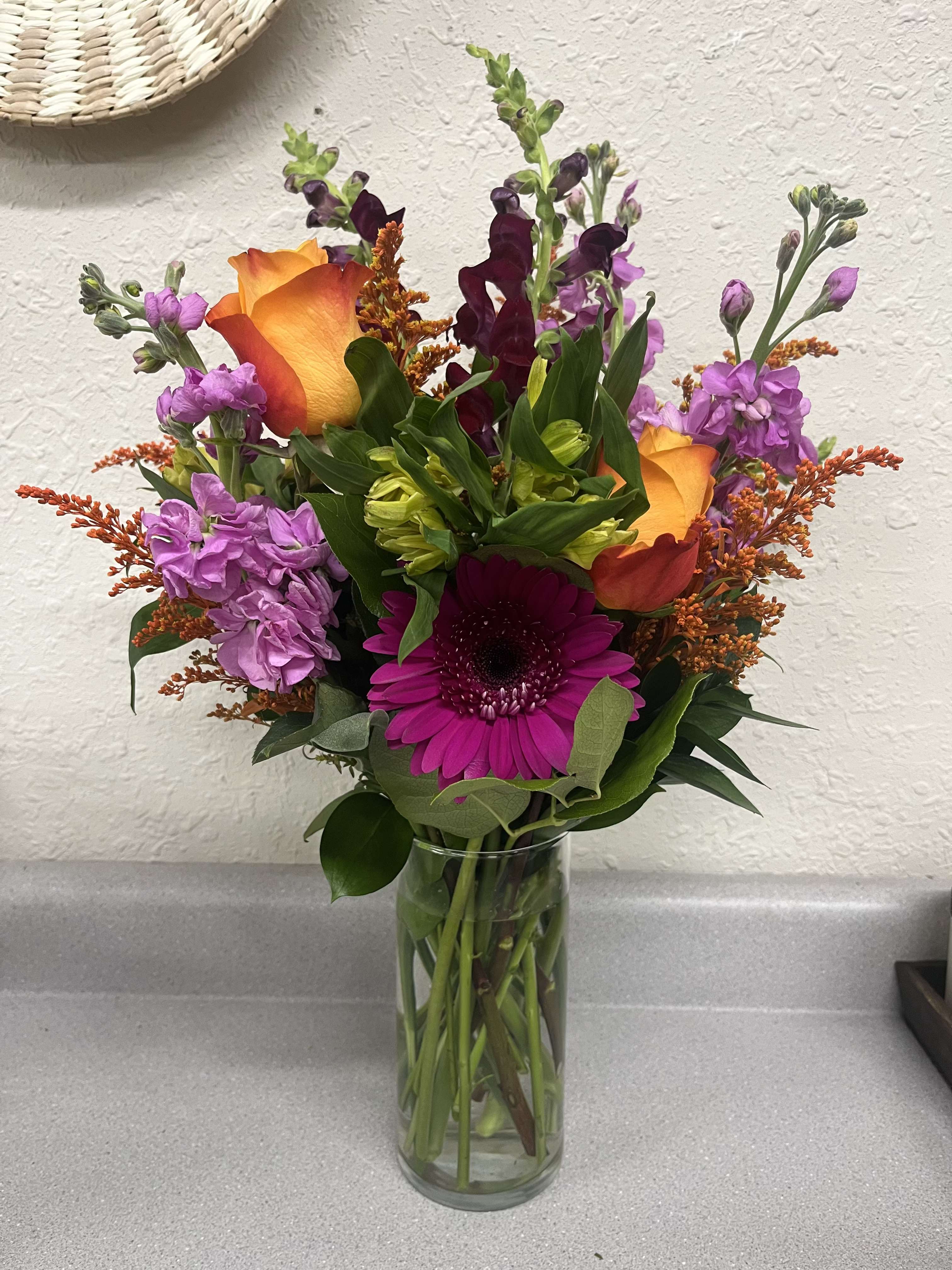 Mixed bouquet with orange roses, purple blooms, and a magenta gerbera in a glass vase