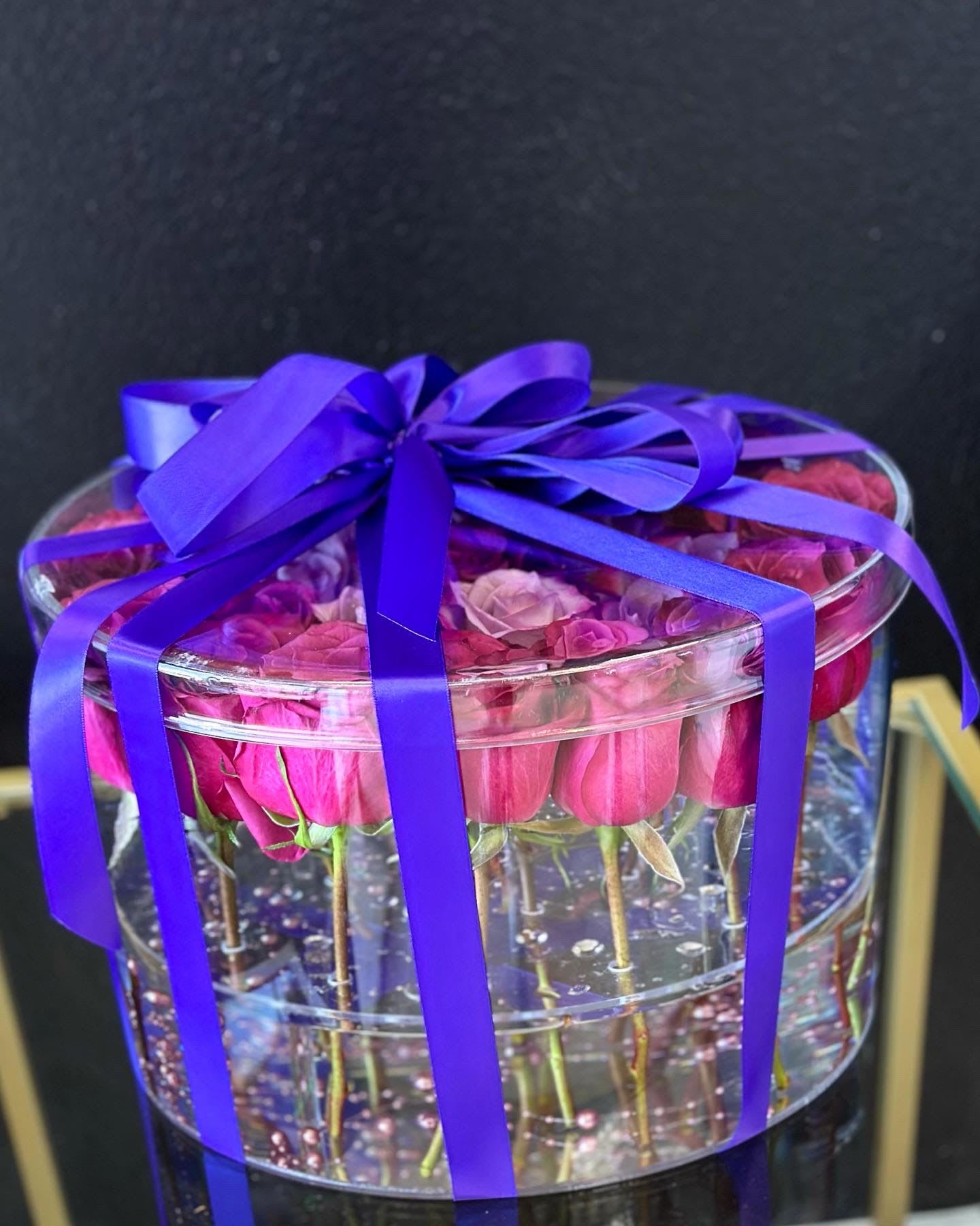 Pink roses in a clear round box tied with a purple ribbon