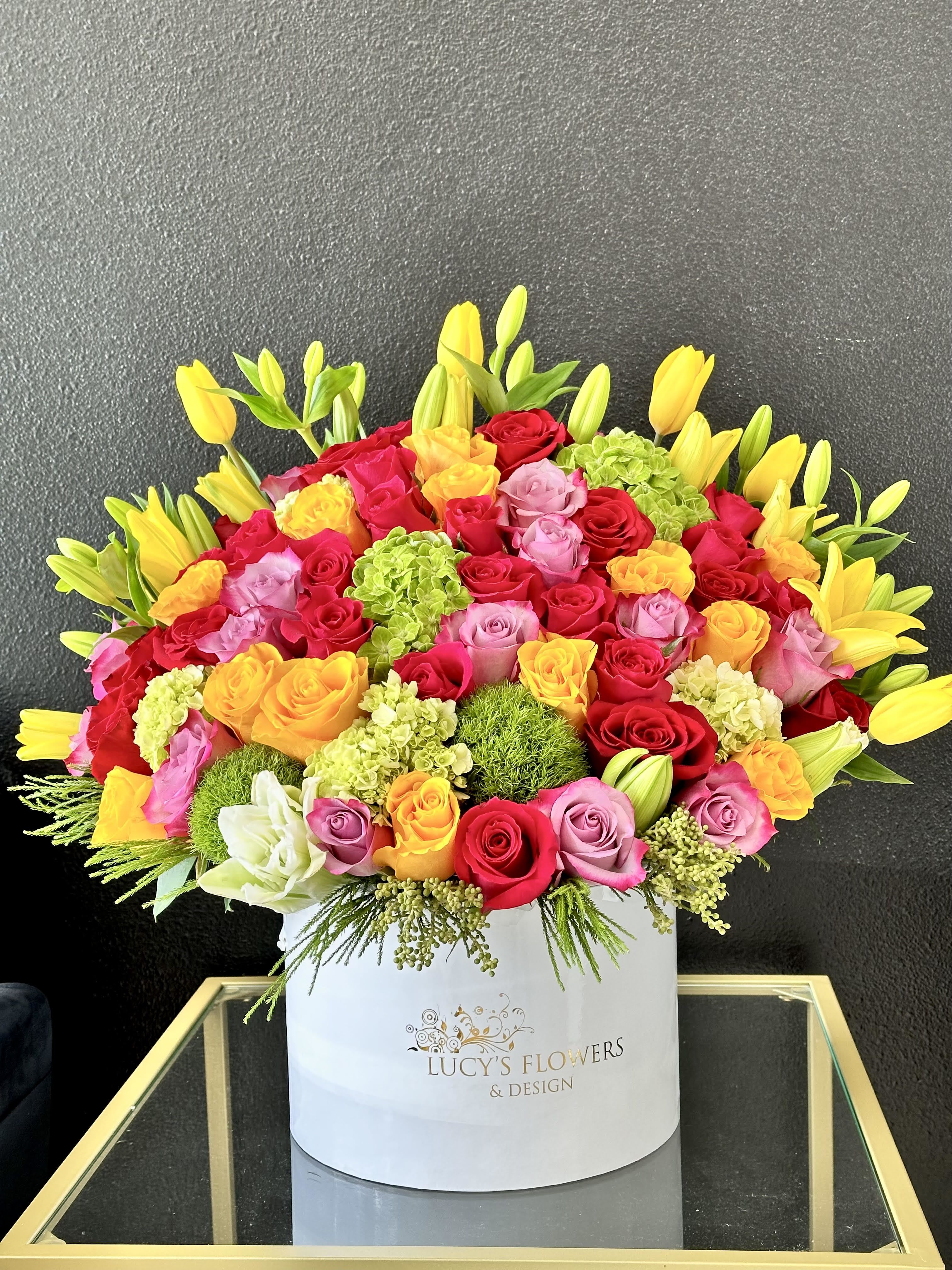 Bright arrangement of multicolor roses, yellow tulips, lilies, and green blooms in a white hat box.