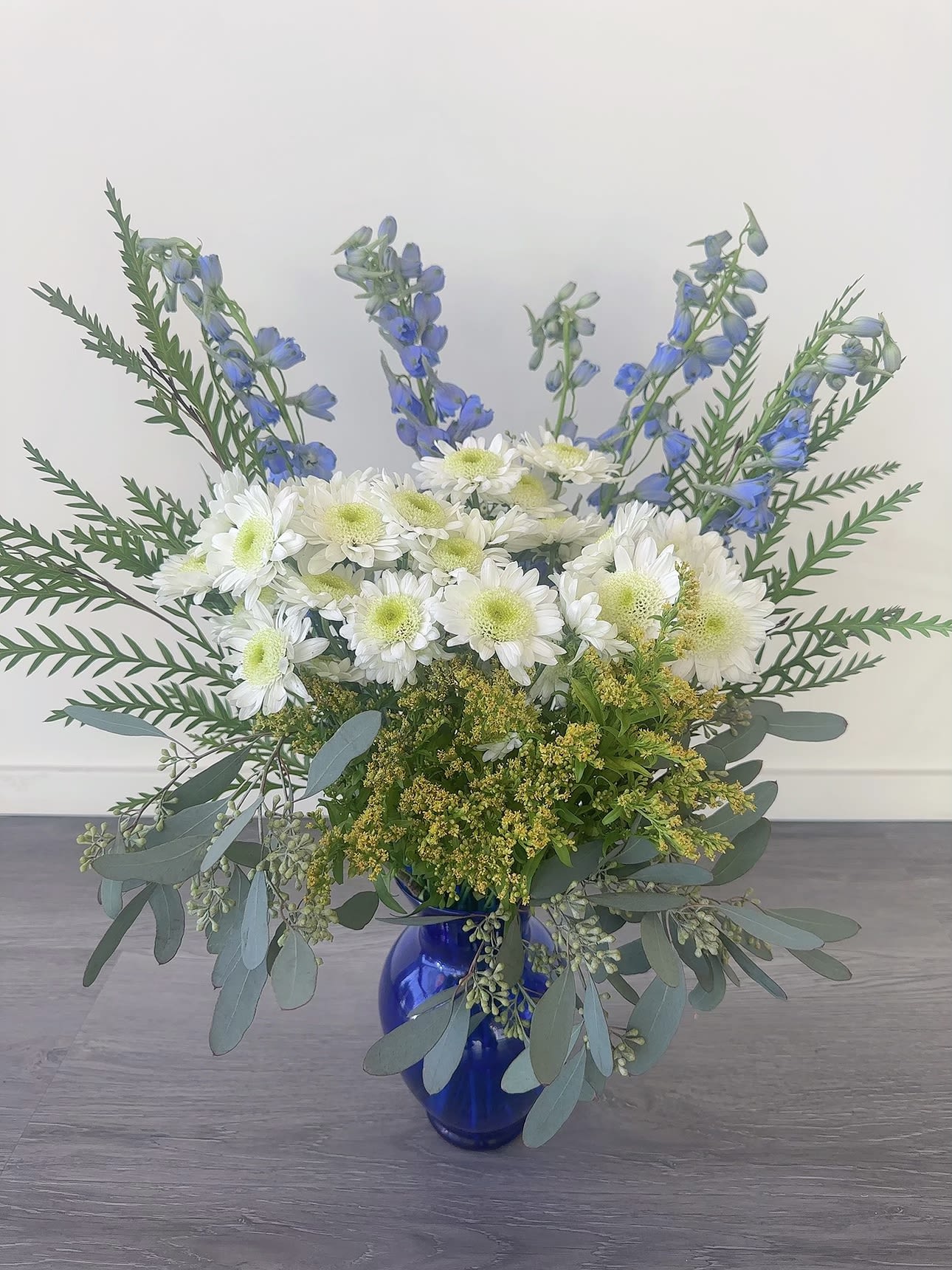 daisy love - This tall blue and white arrangement is a breezy breath of summer air in an interesting vase. Featuring goldenrod, delphinium, and white daisy poms this arrangement is a sweet summer gesture. Send it for birthdays or as a just-because surprise.  Approximate Dimensions: 18"D X 32"H
