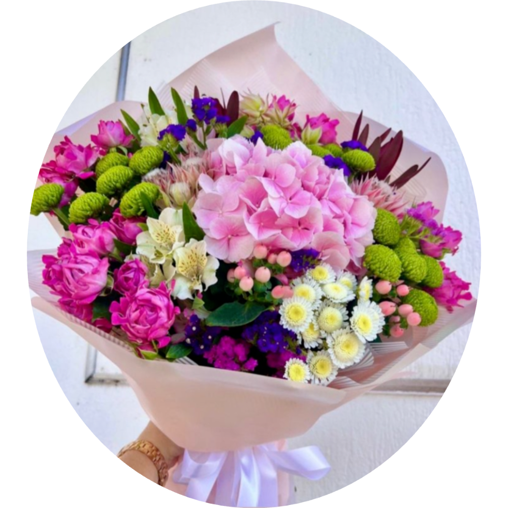 Handheld bouquet of pink, green, white, and purple flowers wrapped in pale paper